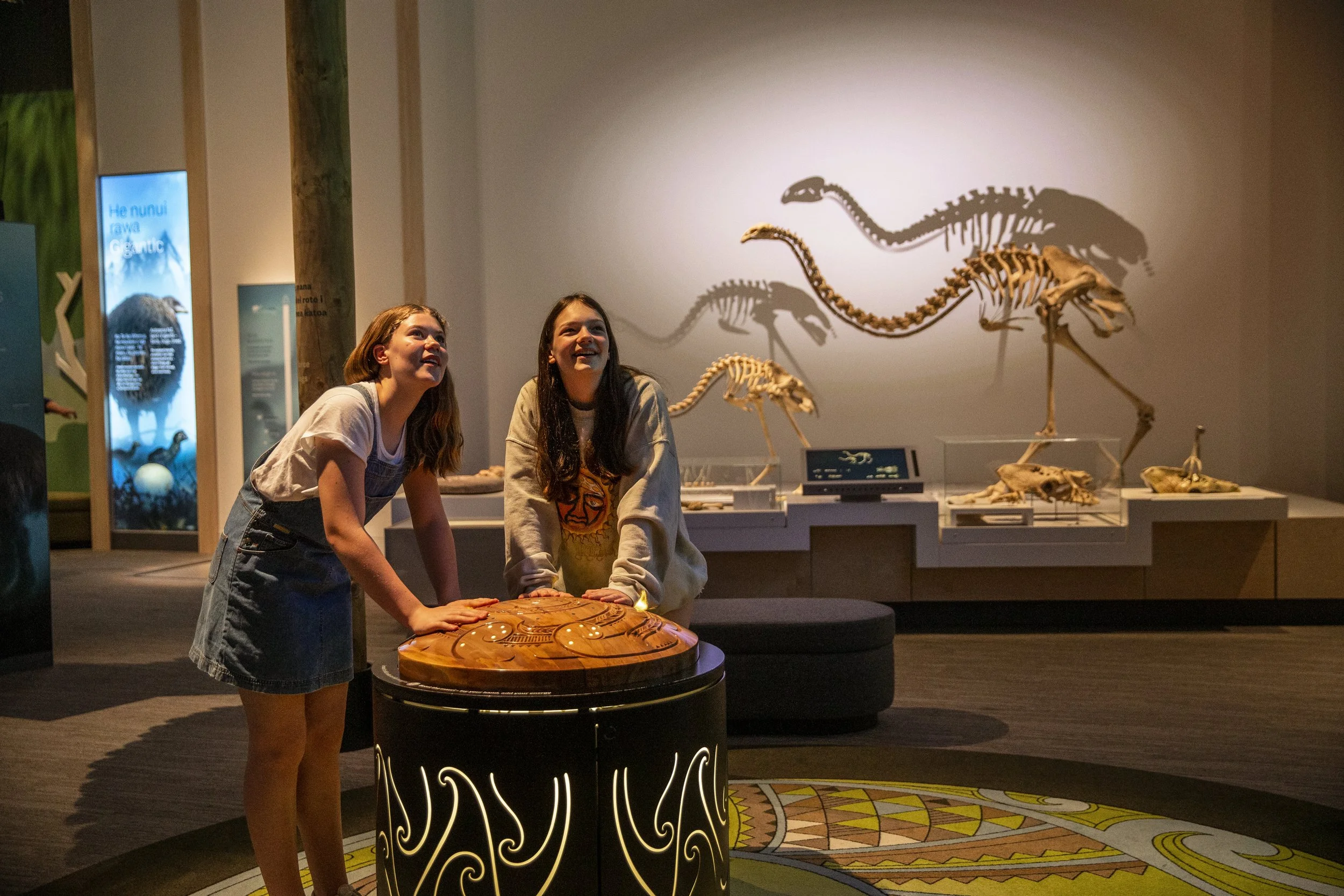 Two young women exploring an interactive exhibit at a museum with large bird skeletons displayed in the background.