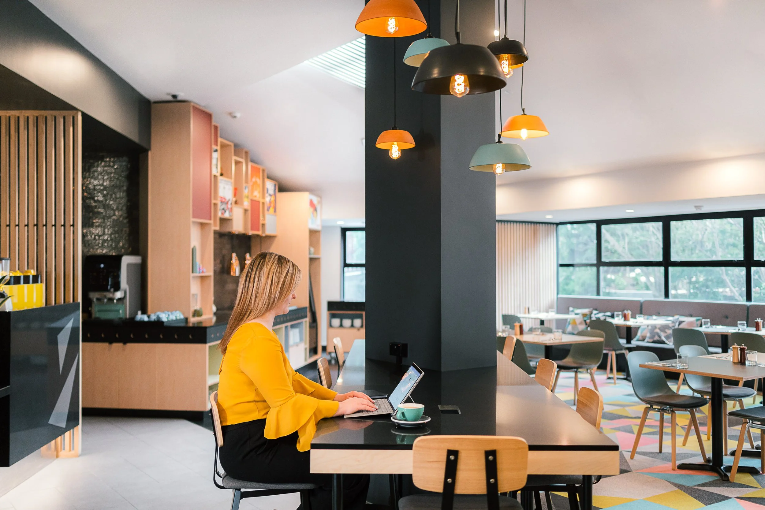 Guest working on a laptop in a modern hotel café with contemporary seating and warm lighting.
