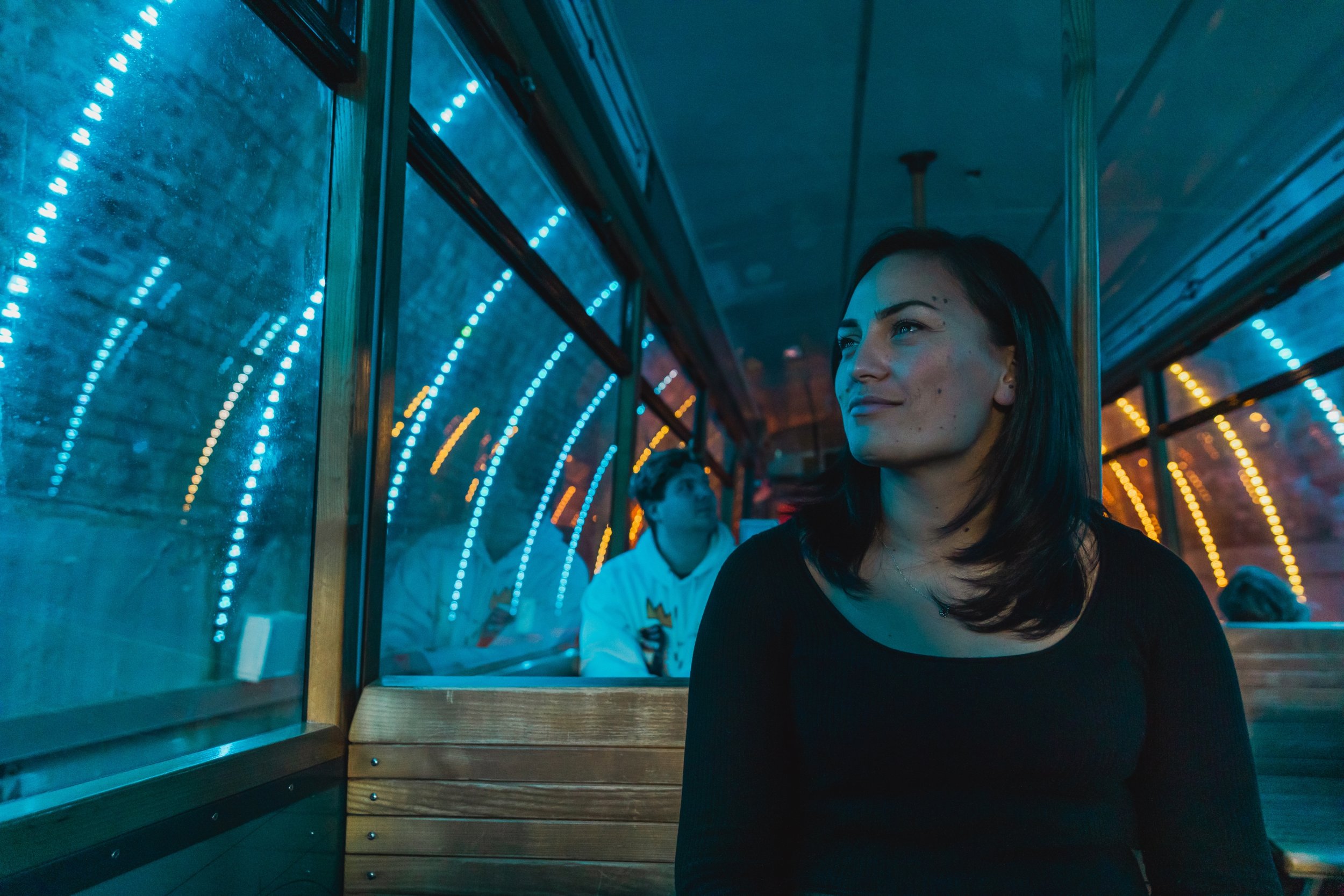 Woman sitting inside a tram at night, illuminated by blue and orange city lights reflecting on the window.
