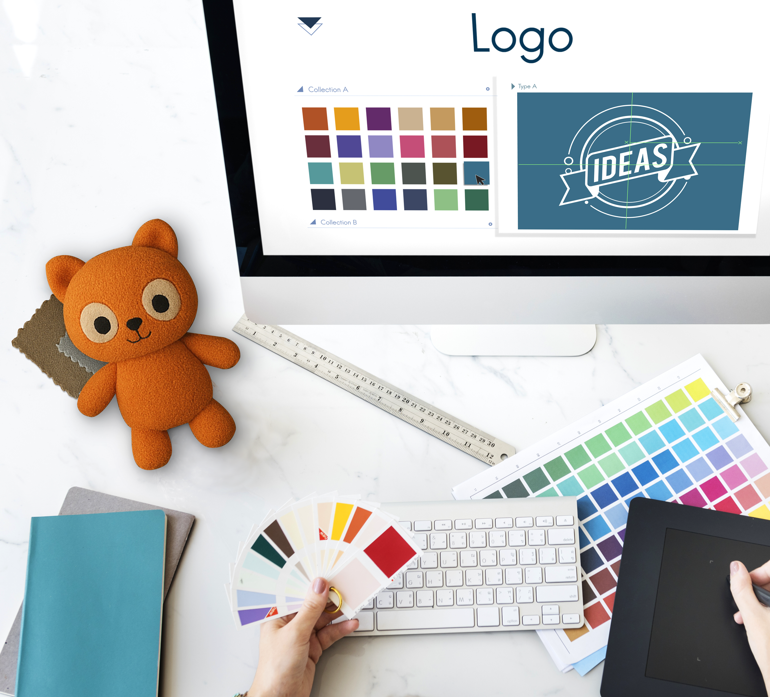 Design workspace with color swatches, a computer screen displaying color palettes and a logo template, a stuffed orange bear toy, a color fan deck, a ruler, notebooks, a keyboard, a graphics tablet with a stylus, and a person's hand holding the color fan.