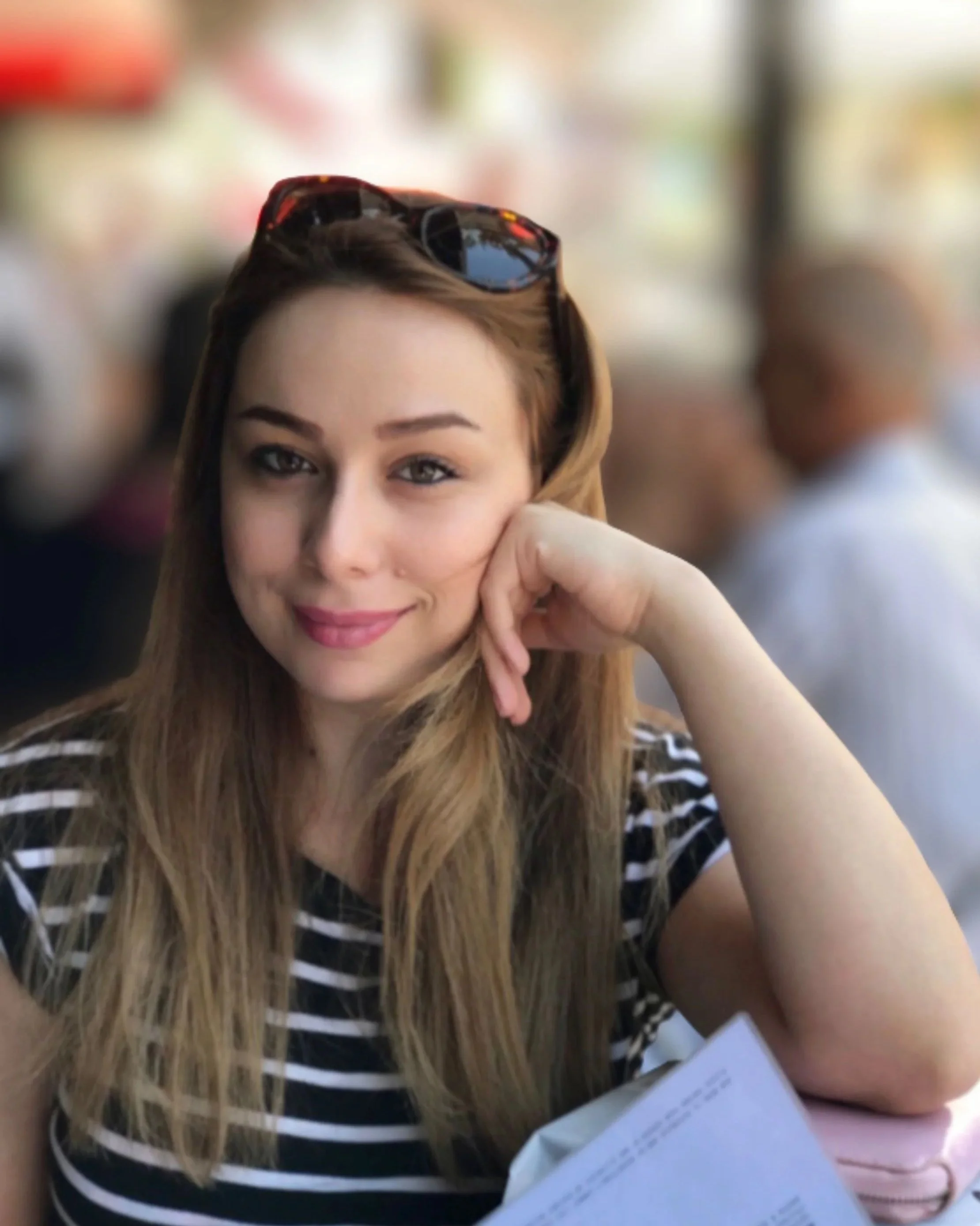 A woman with long, light brown hair, wearing a black and white striped shirt, sitting with her hand resting on her face, sunglasses on her head, smiling softly, with a blurred background of people and colorful objects.