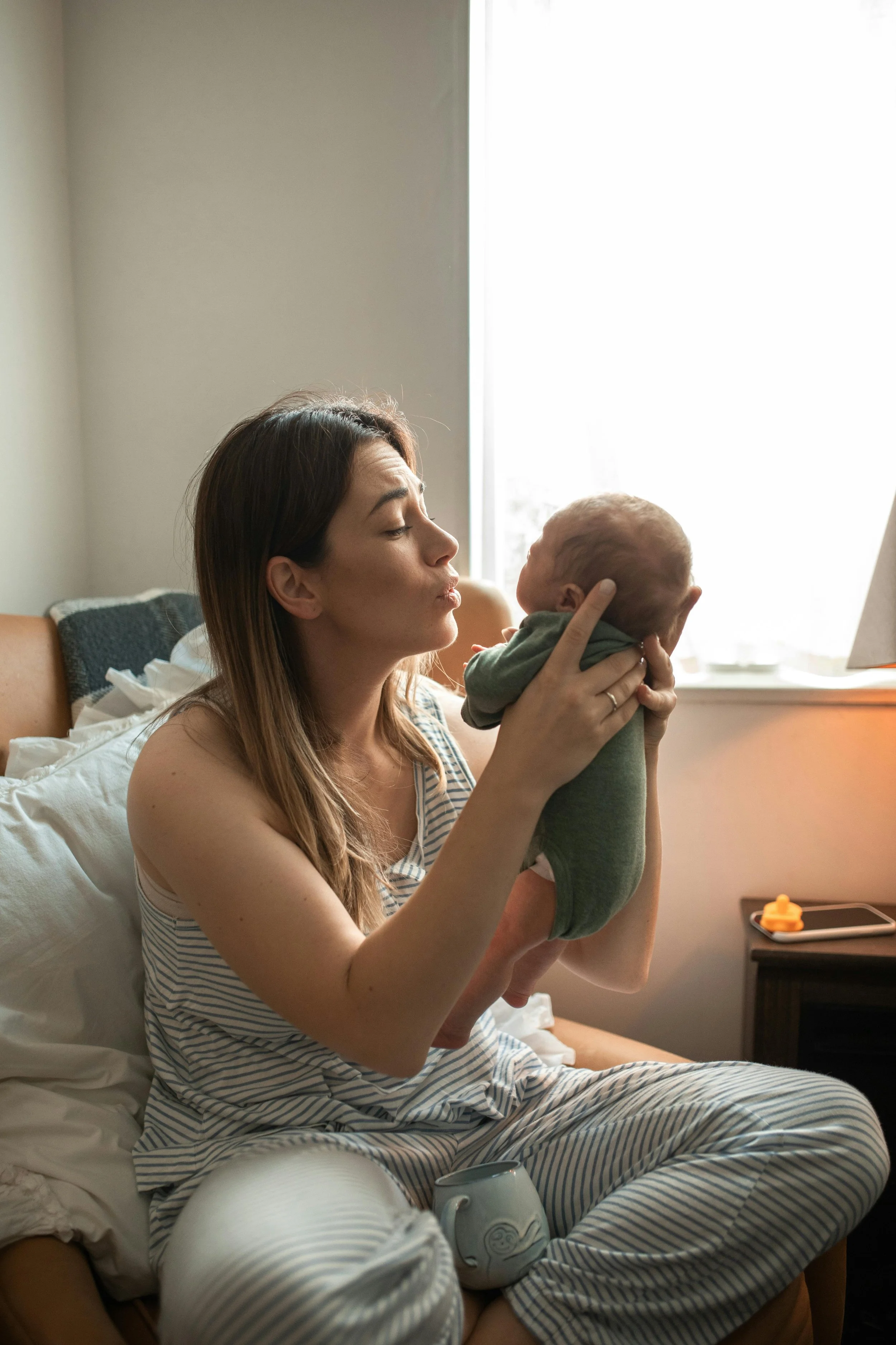 A woman in pajamas holding a baby in a room with natural light. The woman is sitting on a bed and gently holding the baby up, with her eyes closed and a slight smile.