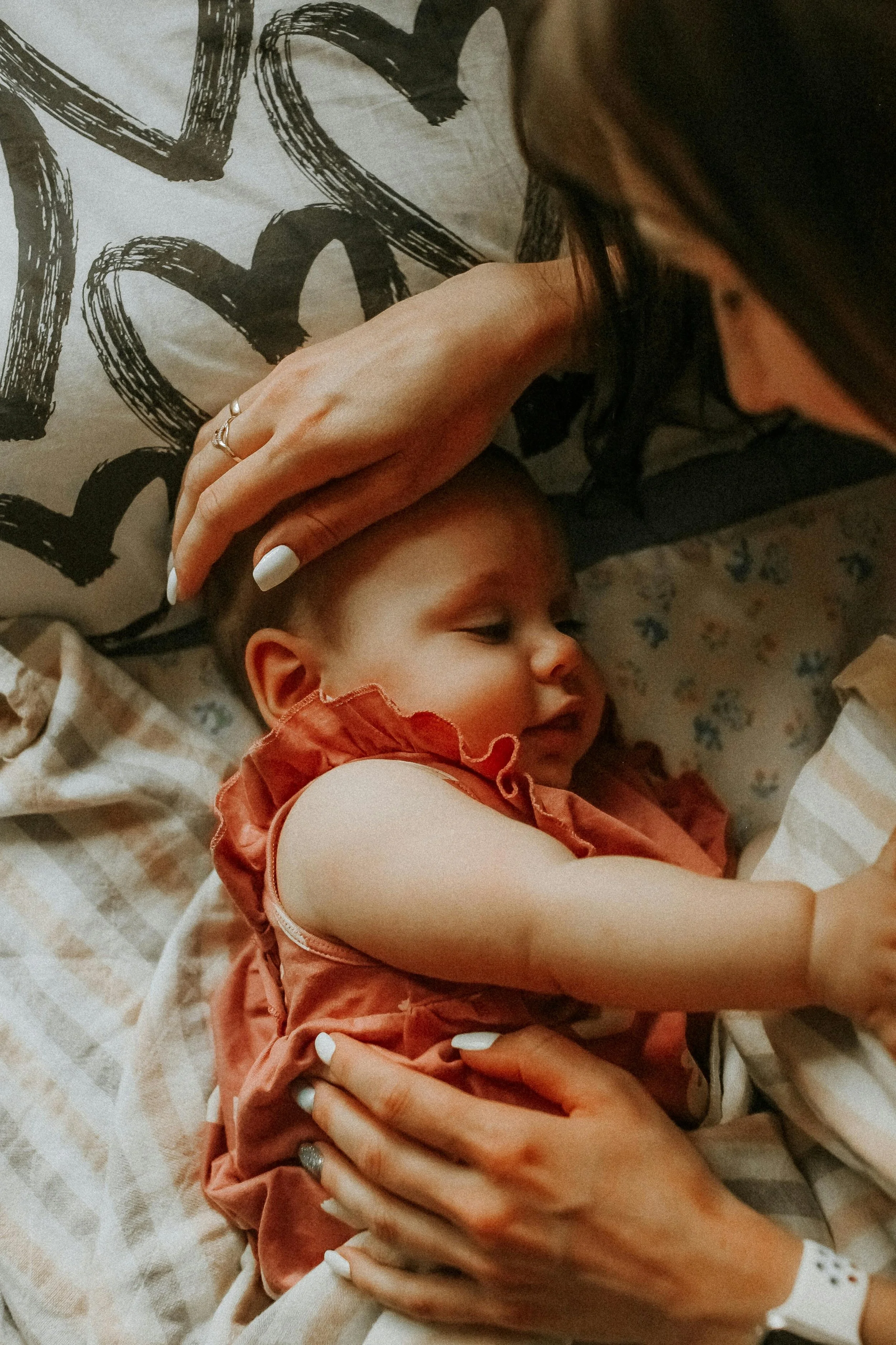 A woman gently touches a sleeping baby lying on a bed, with the woman having manicured nails and multiple rings on her fingers. The baby is wearing a pinkish-brown dress and appears relaxed.
