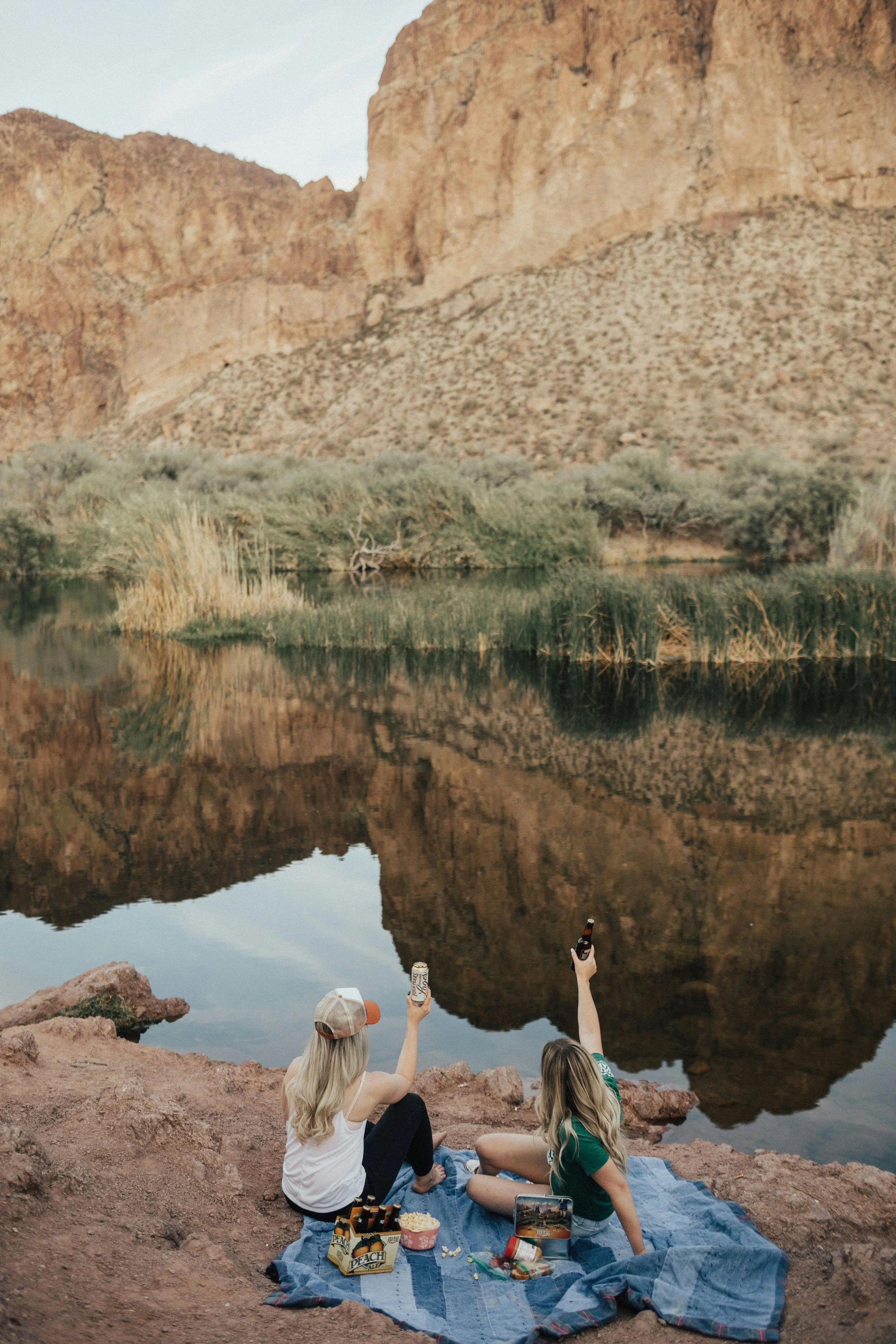 Two women sitting on a blanket by a river, with mountains in the background, holding drinks and enjoying a picnic.