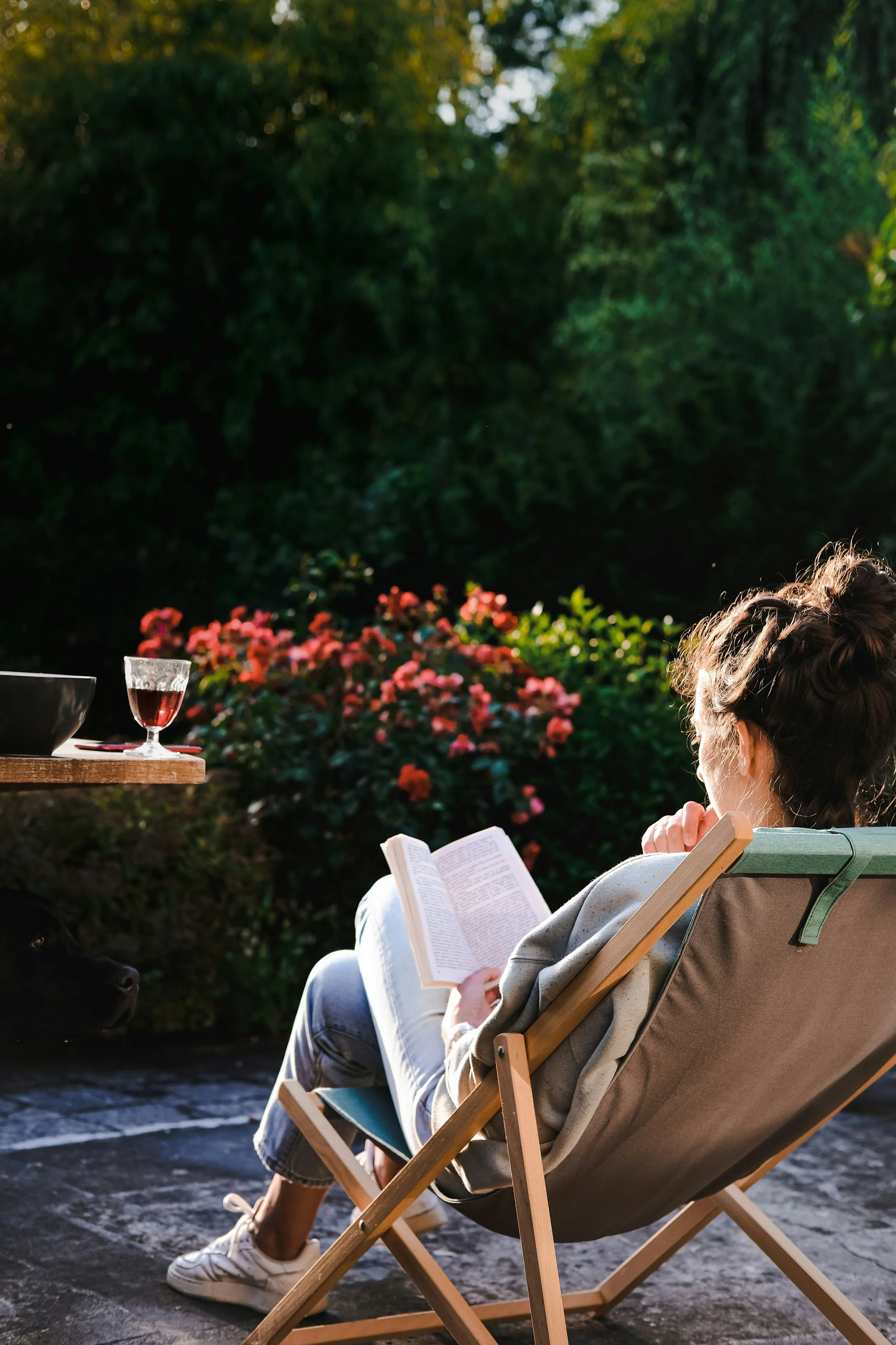 A woman sitting in a deck chair outdoors, reading a book, with a glass of red wine on a table and a dog nearby. In the background, there are blooming bushes and trees.