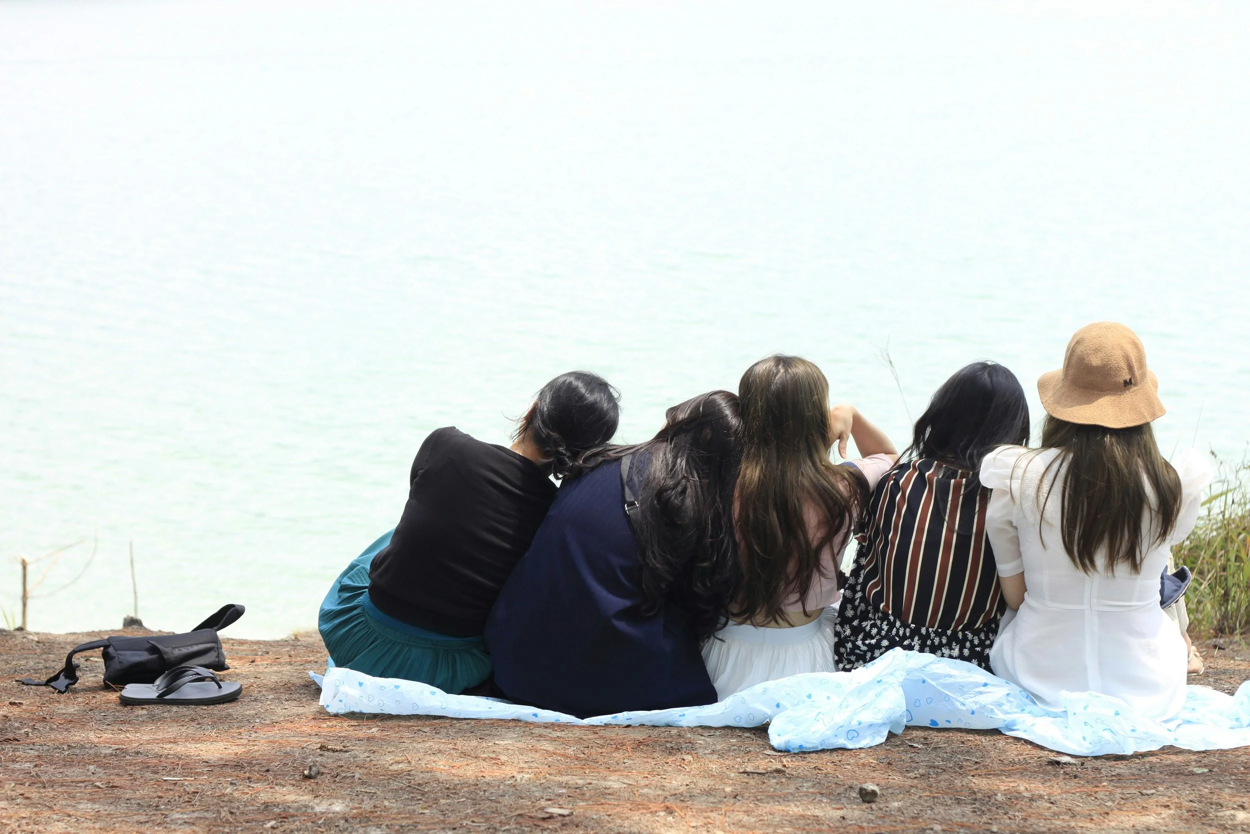 Five women sitting on a blanket near a body of water, viewed from the back, with some wearing hats and one with long dark hair, during daytime.