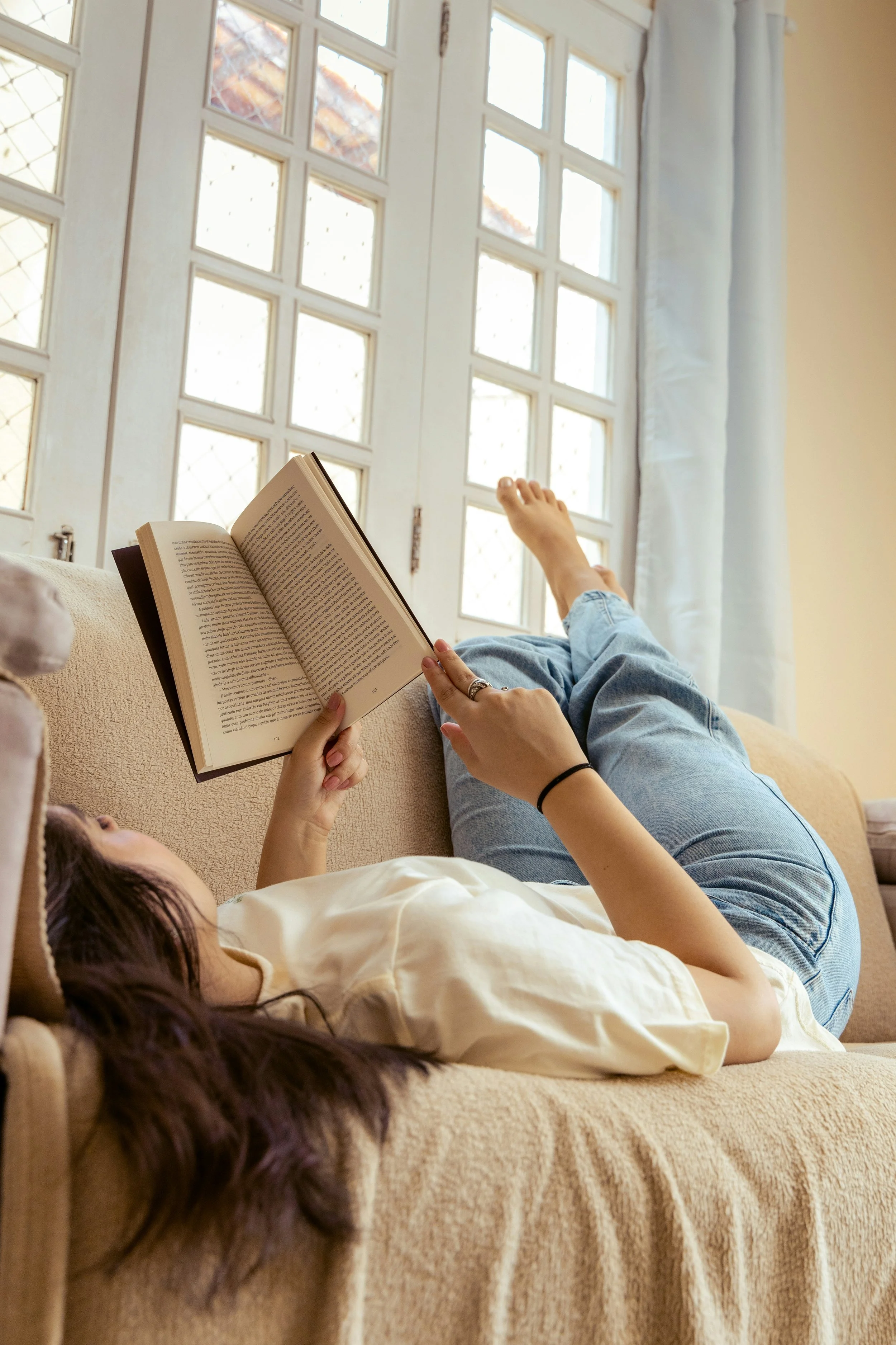 A person lying on a beige sofa, reading a book, with their legs stretched out, next to a glass-paneled white door and white curtains.