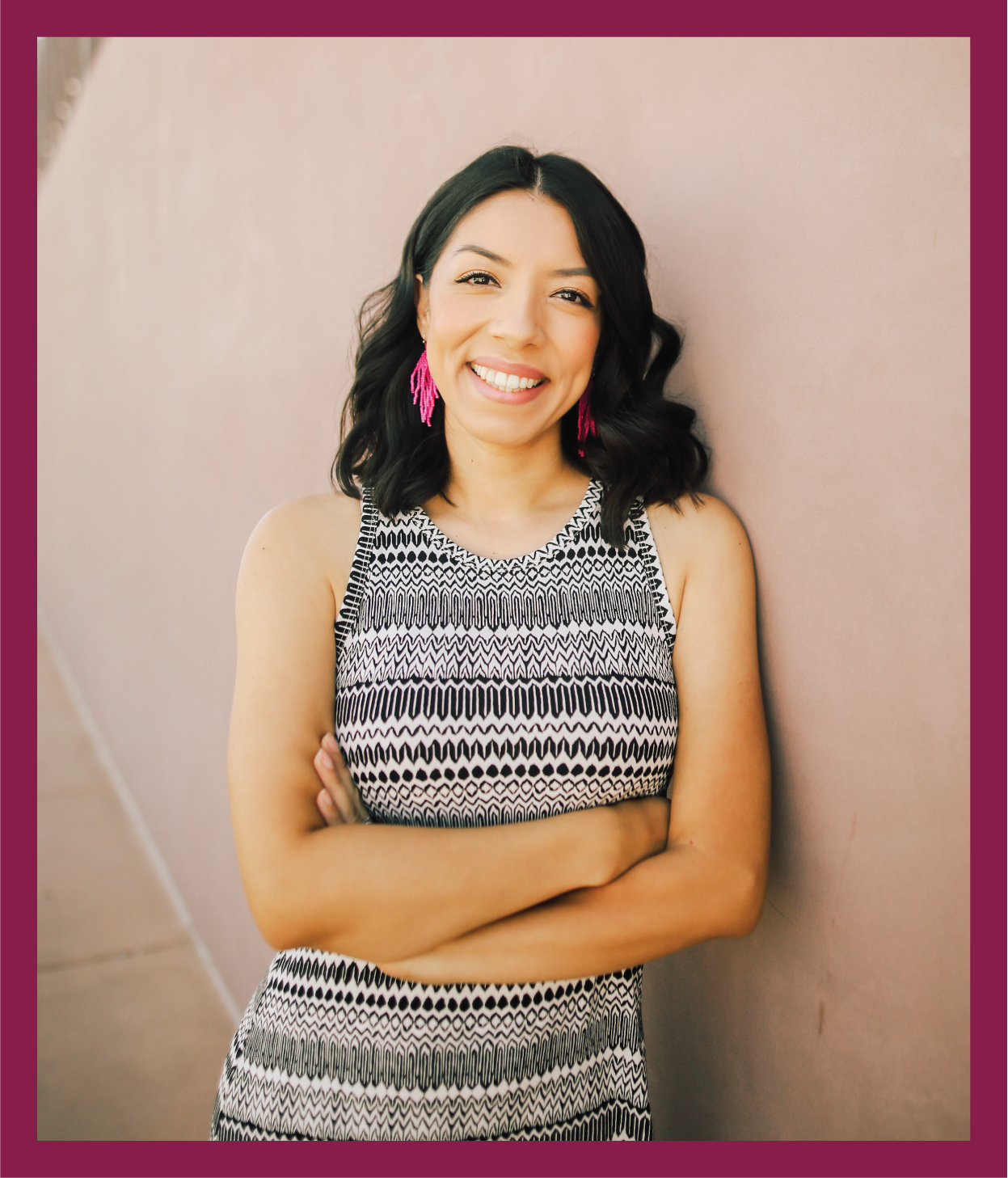 A woman with shoulder-length black hair, wearing pink earrings and a sleeveless black and white patterned dress, smiling and standing with arms crossed against a pink wall.