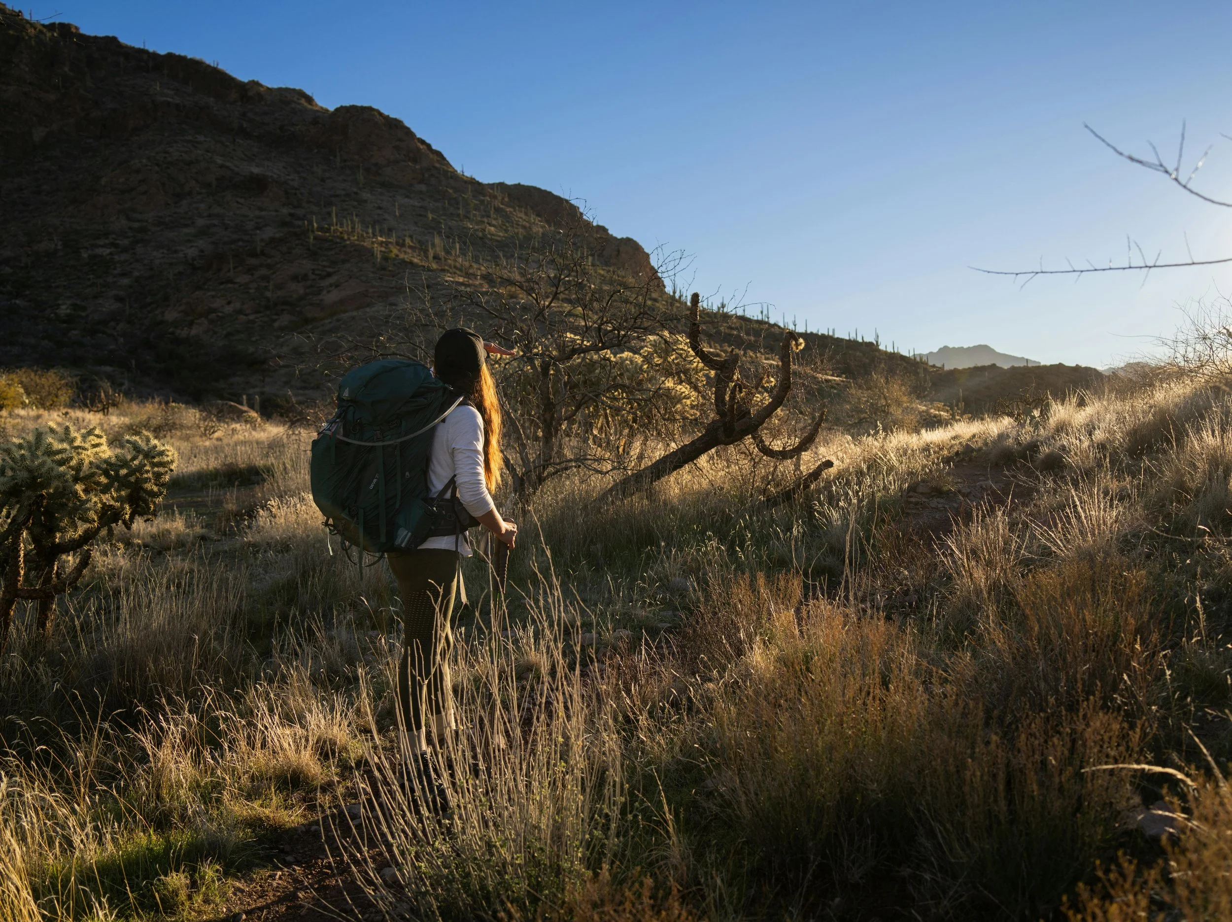 A woman with a backpack hiking in a desert landscape during sunrise or sunset, with mountains and cacti in the background.