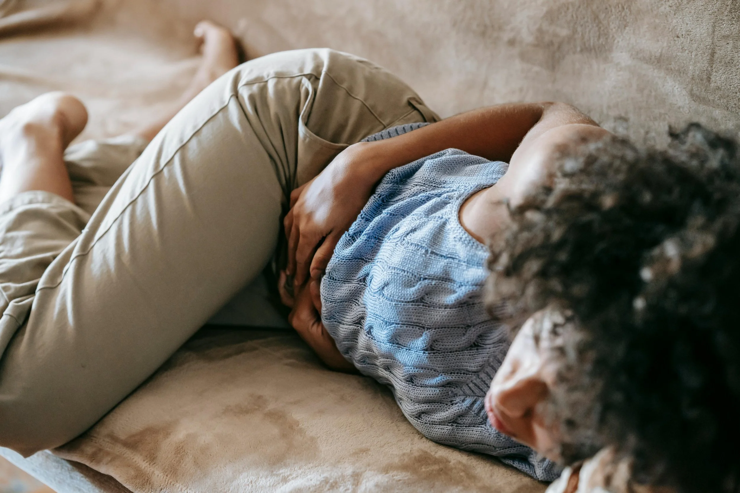 Person lying on a couch with their arm over their face, wearing a blue sweater and beige pants.