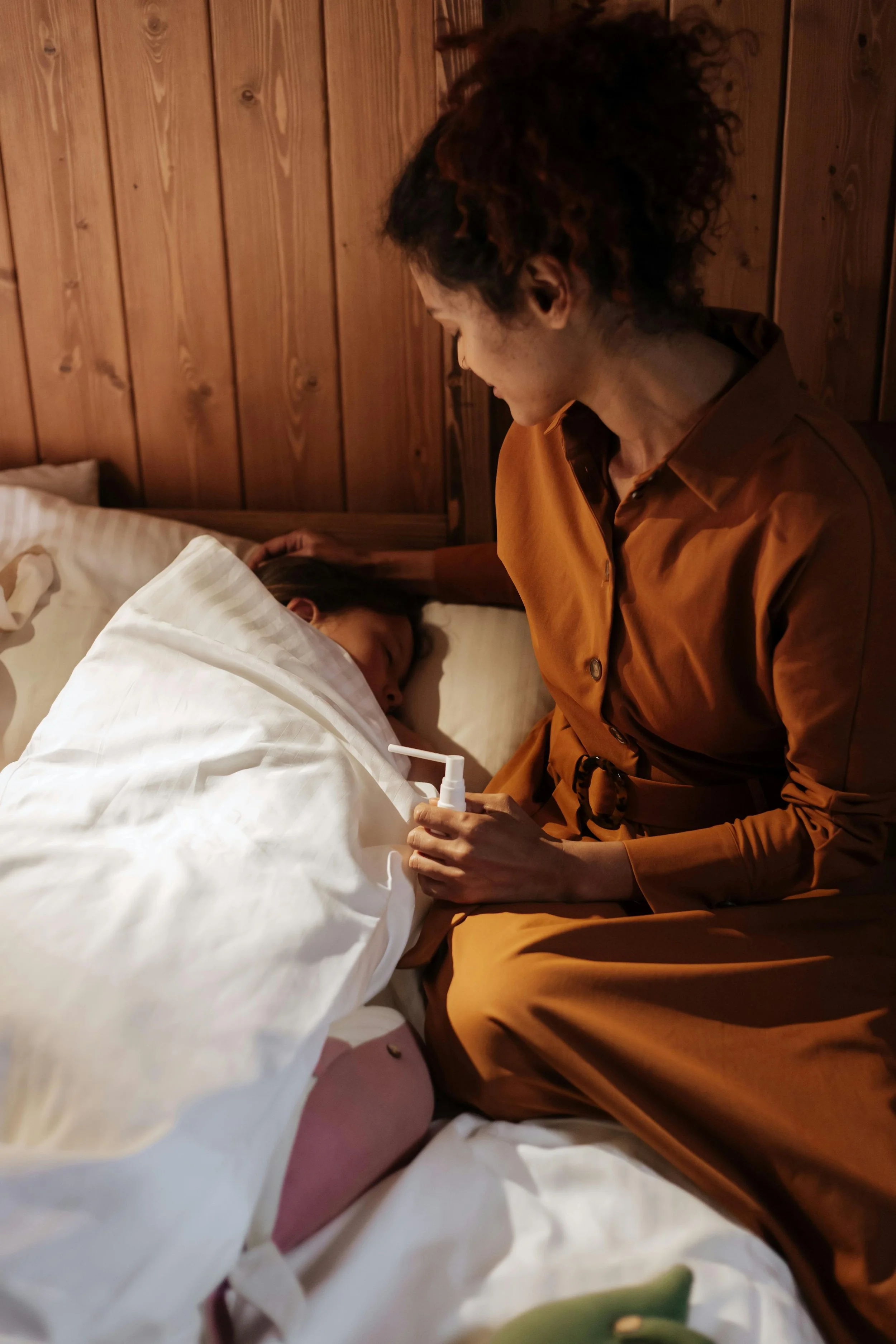 A woman with curly hair wearing a brown dress, sitting on a bed and holding a nasal spray bottle, looking at a woman lying in bed with her eyes closed.