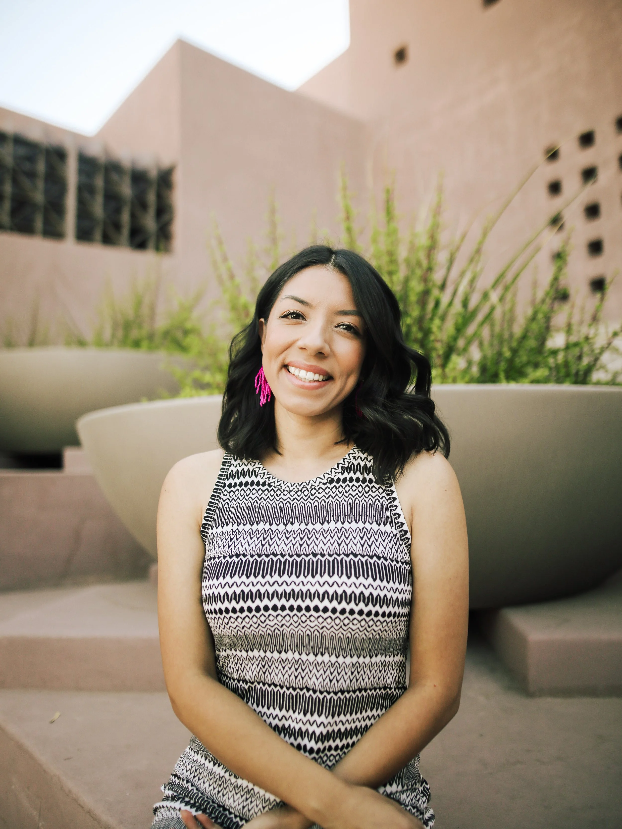 A young woman with black wavy hair, wearing pink earrings and a sleeveless patterned dress, smiling while sitting outdoors in front of large beige planters with green plants and pinkish building structures in the background.