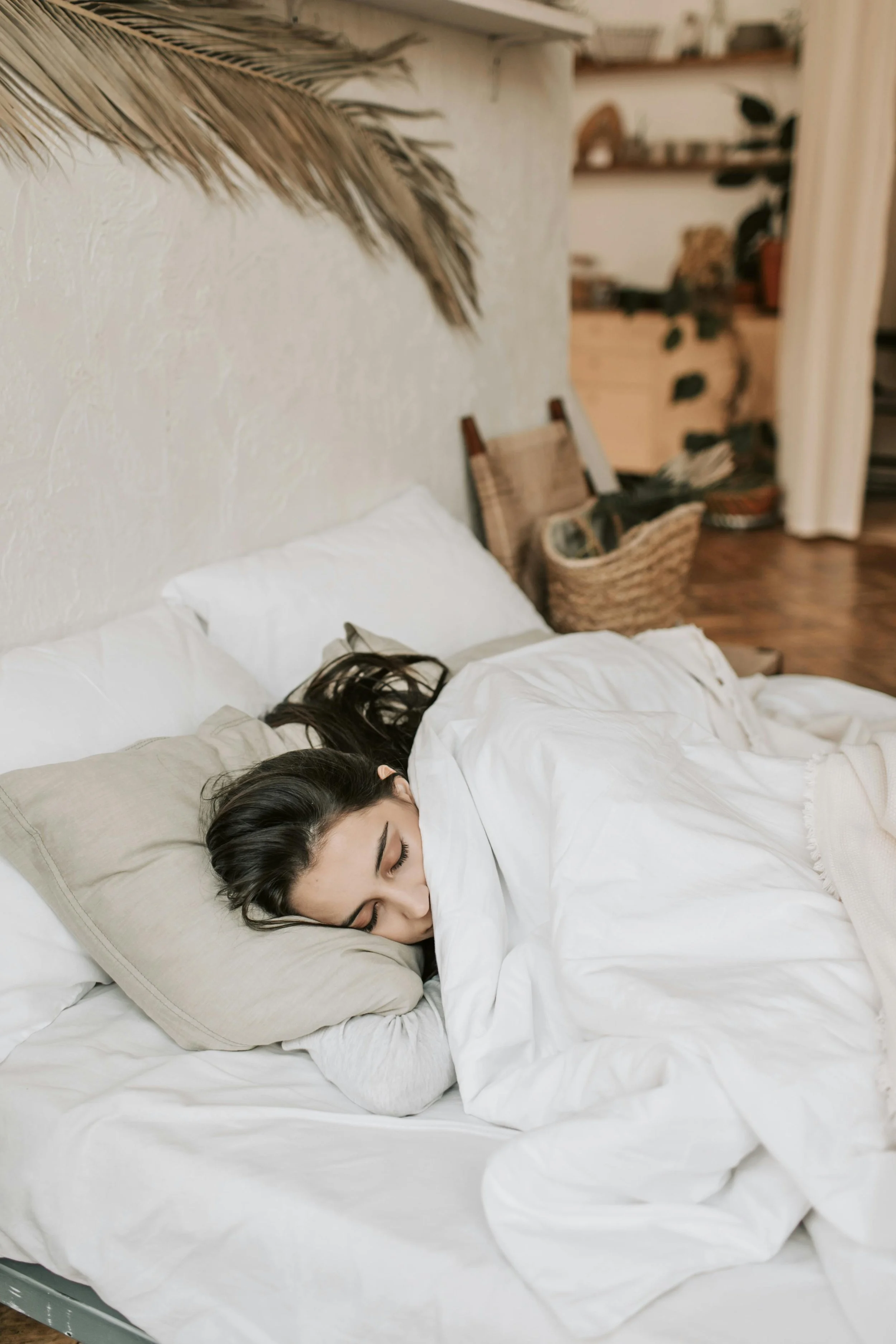 A woman sleeping on her side in a bed with white sheets and beige pillows in a cozy room with wooden floors and a wicker basket in the background.