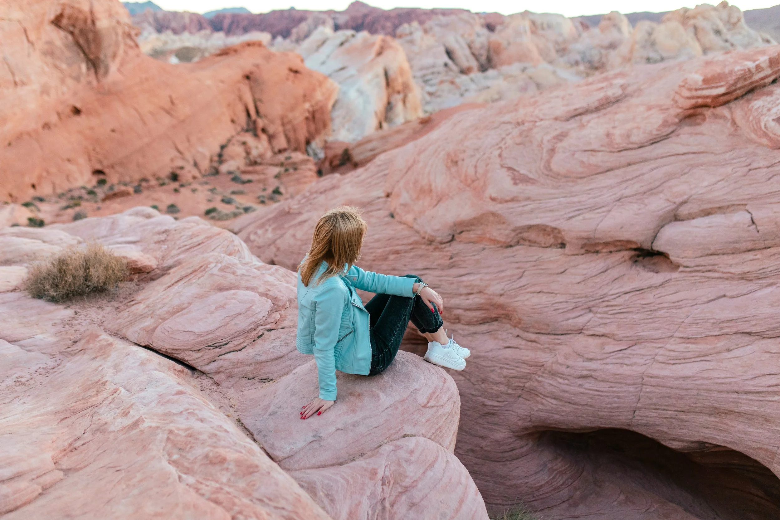 A woman sitting on a pinkish rock formation in a desert landscape with multicolored hills in the background.