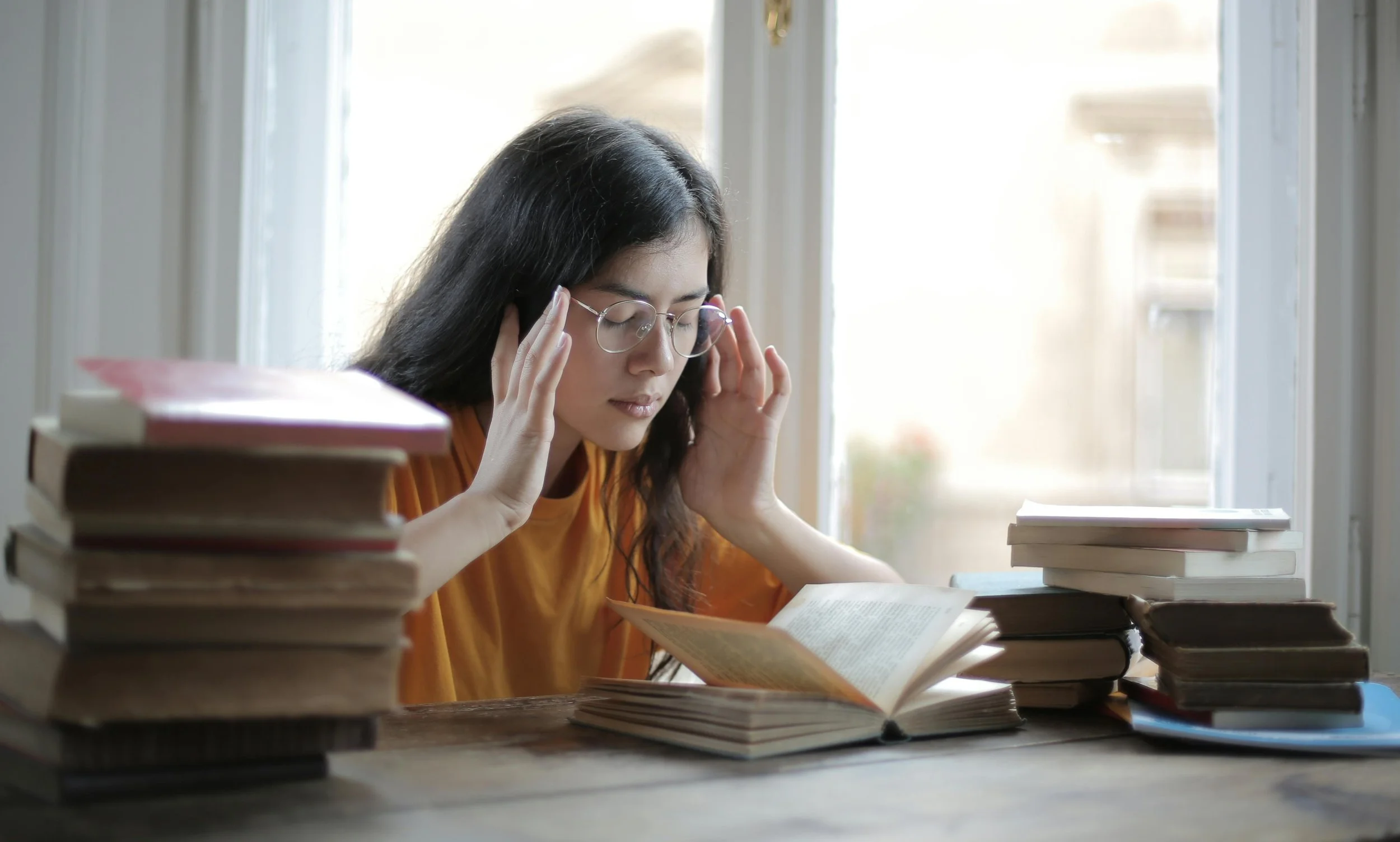 A young woman with glasses is sitting at a table surrounded by stacks of books. She is holding her head with both hands while reading a book in front of her, with a window in the background letting in natural light.