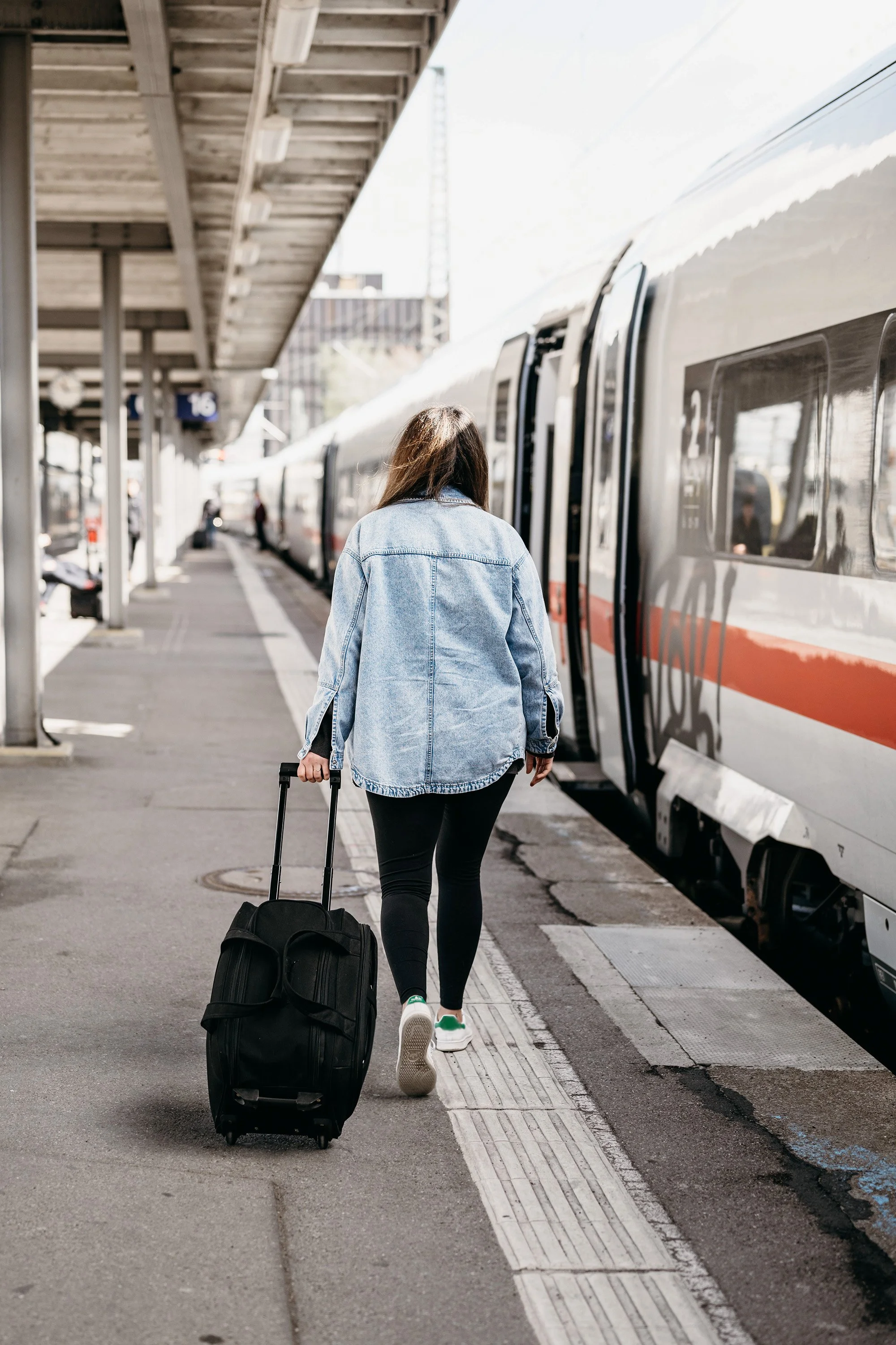 A woman with long brown hair, wearing a denim jacket, black pants, and white sneakers, pulling a black rolling suitcase as she walks along a train platform beside a parked Amtrak train.