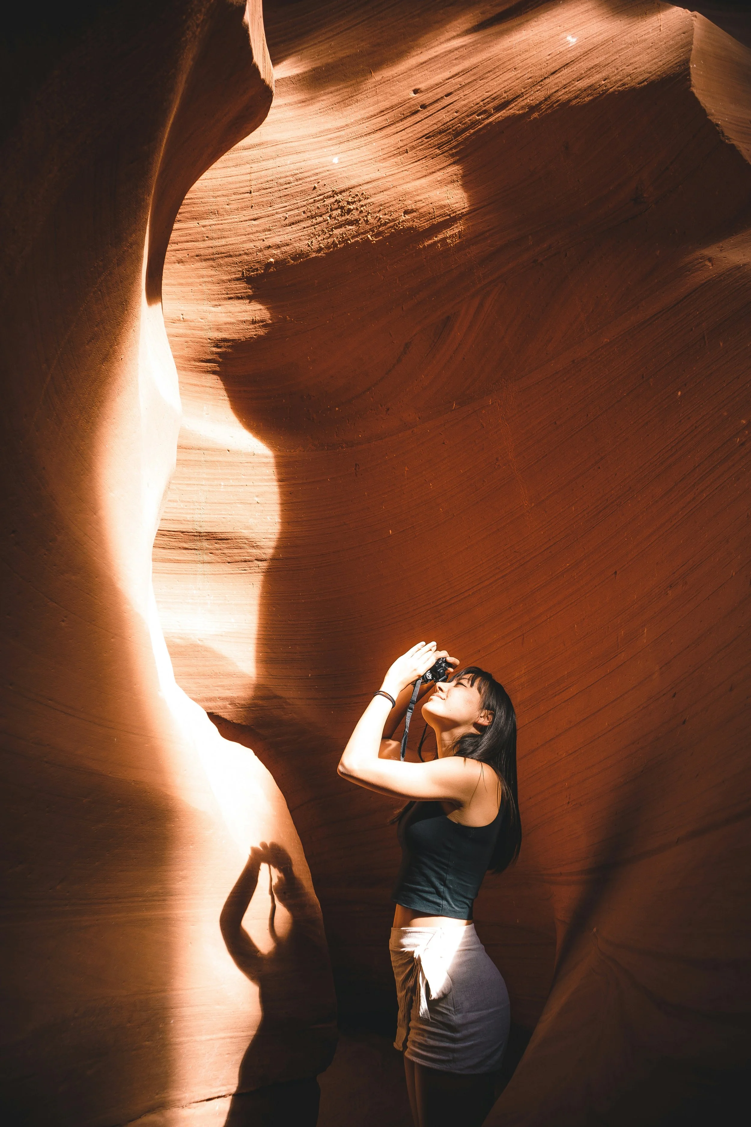 A woman taking a photograph inside a narrow, curved slot canyon with sandstone walls. Sunlight creates shadows and highlights on the rock formations.