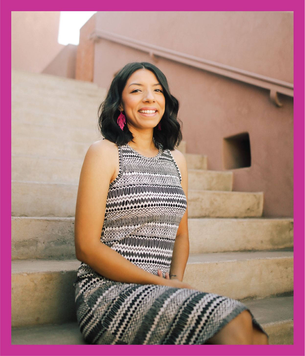 A woman with shoulder-length black hair sitting on outdoor concrete stairs, smiling, wearing a sleeveless patterned dress and pink earrings, with a beige wall in the background.