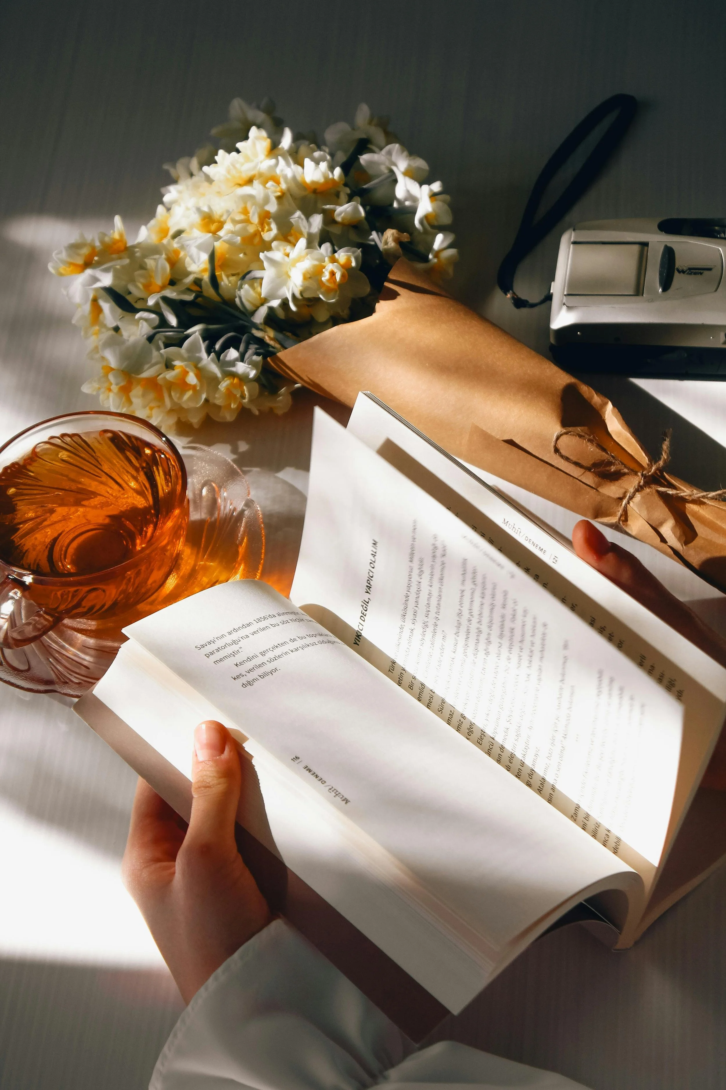 A person holding an open book on a table with a cup of tea, a bouquet of white and yellow flowers, a wrapped gift, and a camera nearby.