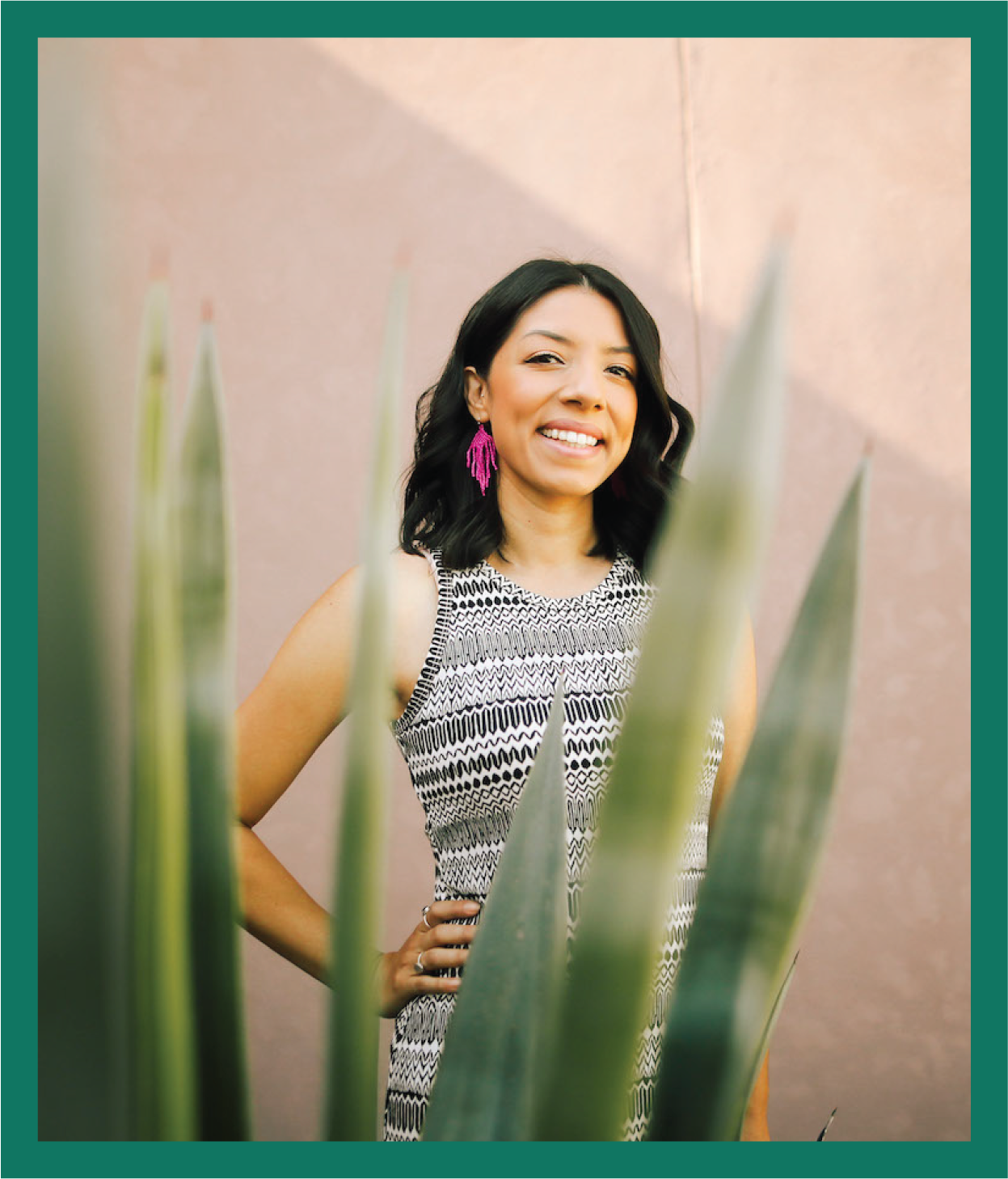 A smiling woman with black hair wearing a patterned sleeveless dress and pink earrings, standing outdoors behind a large green plant with a plain pink background.