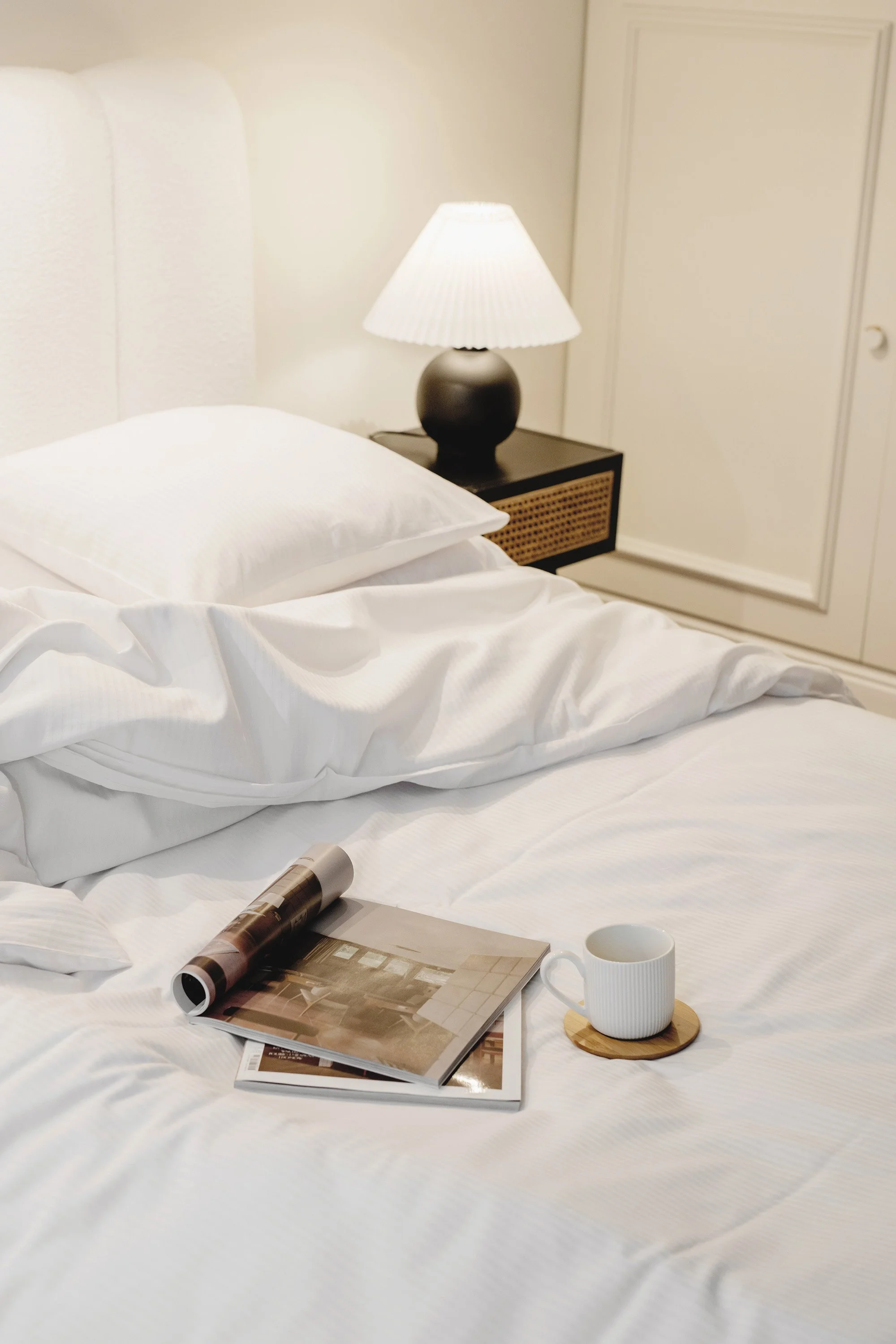 A neatly made bed with white linens, a rolled-up magazine, an open magazine, a white coffee mug on a small wooden coaster, a nightstand with a black lamp on it, and a closed closet door in the background.