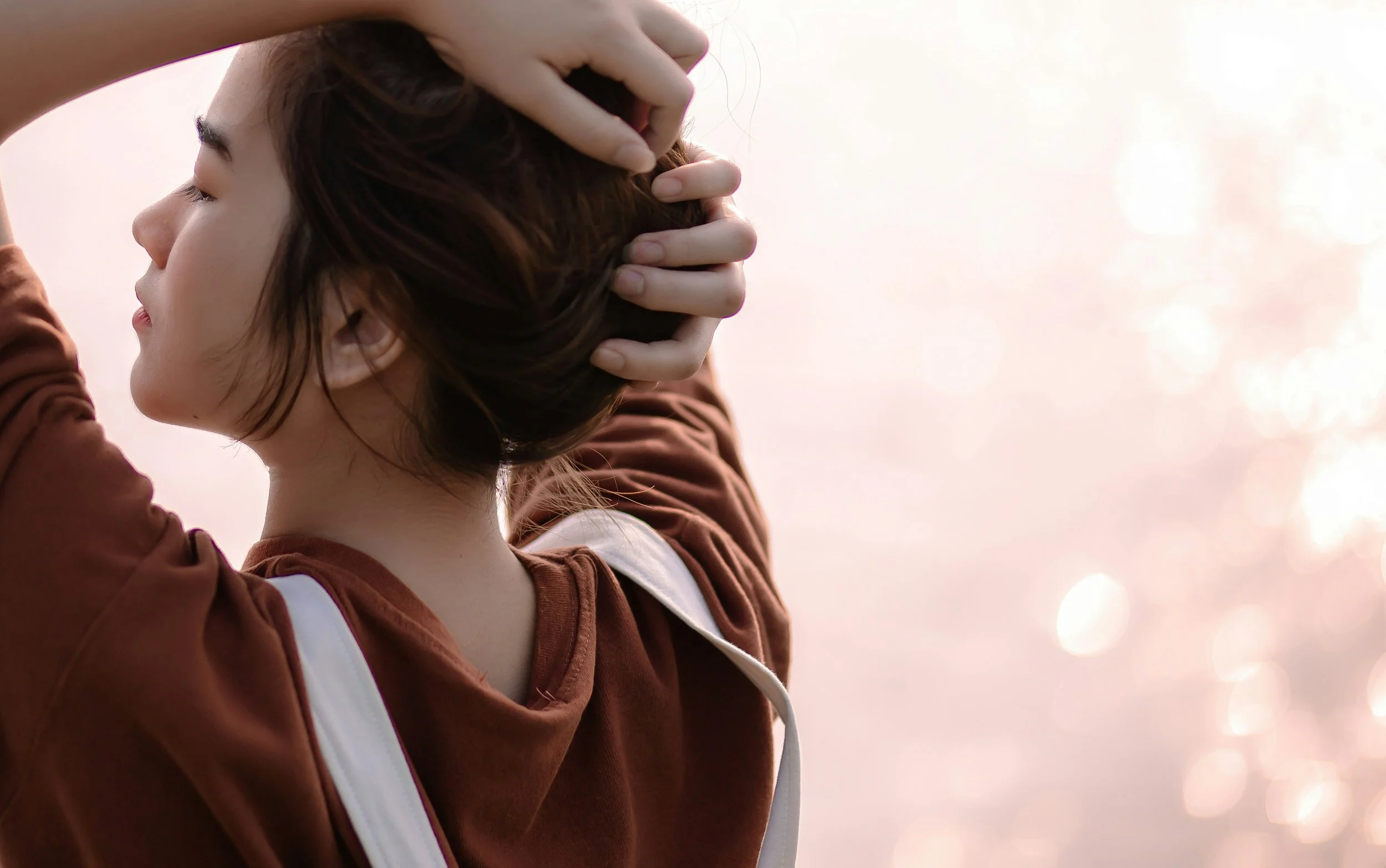 A woman with short brown hair and light skin is standing outdoors, with her eyes closed, holding her head with both hands, against a soft, blurred sunset background.