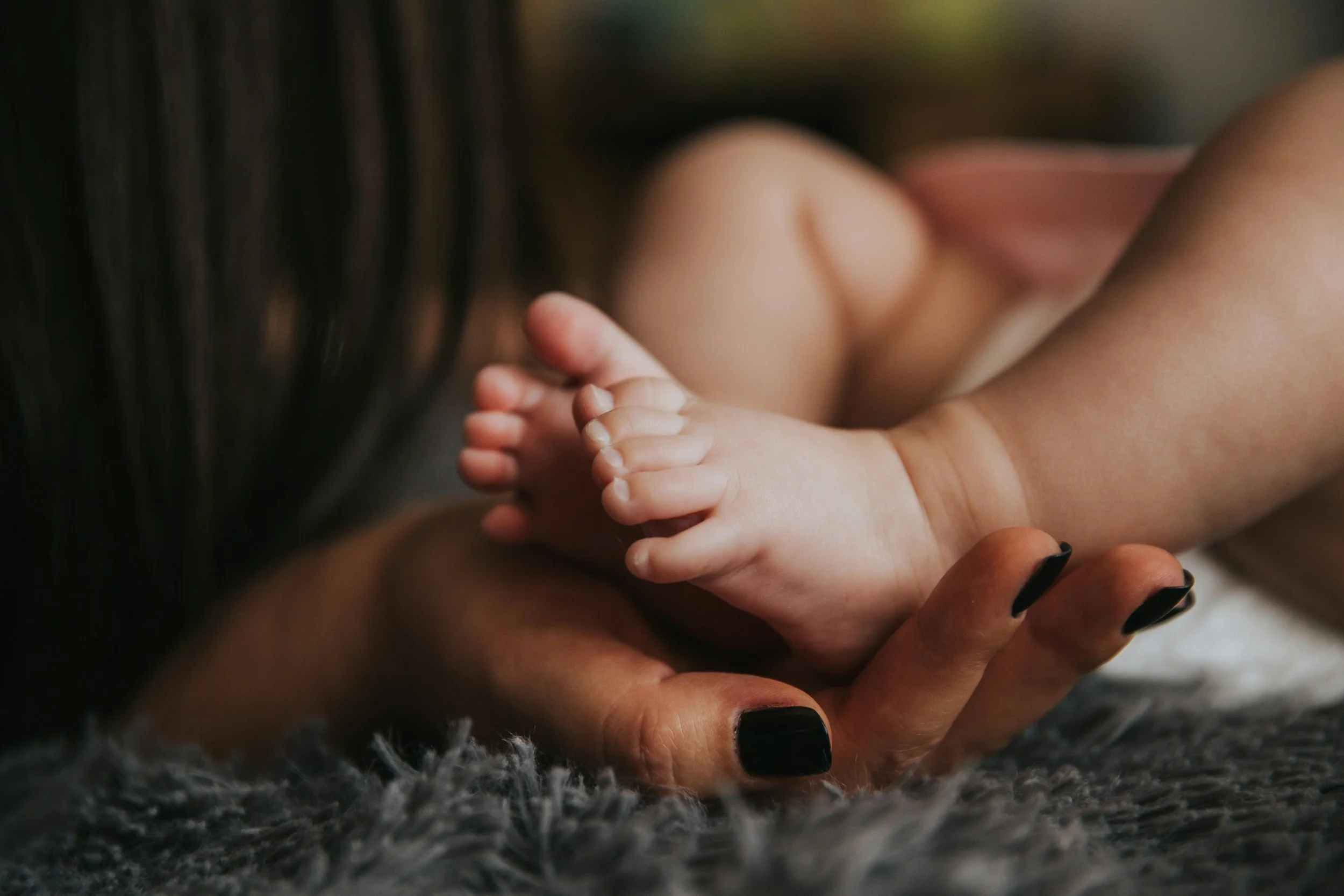 Close-up of a person with painted black nails holding a small baby's feet, lying on a textured gray surface.