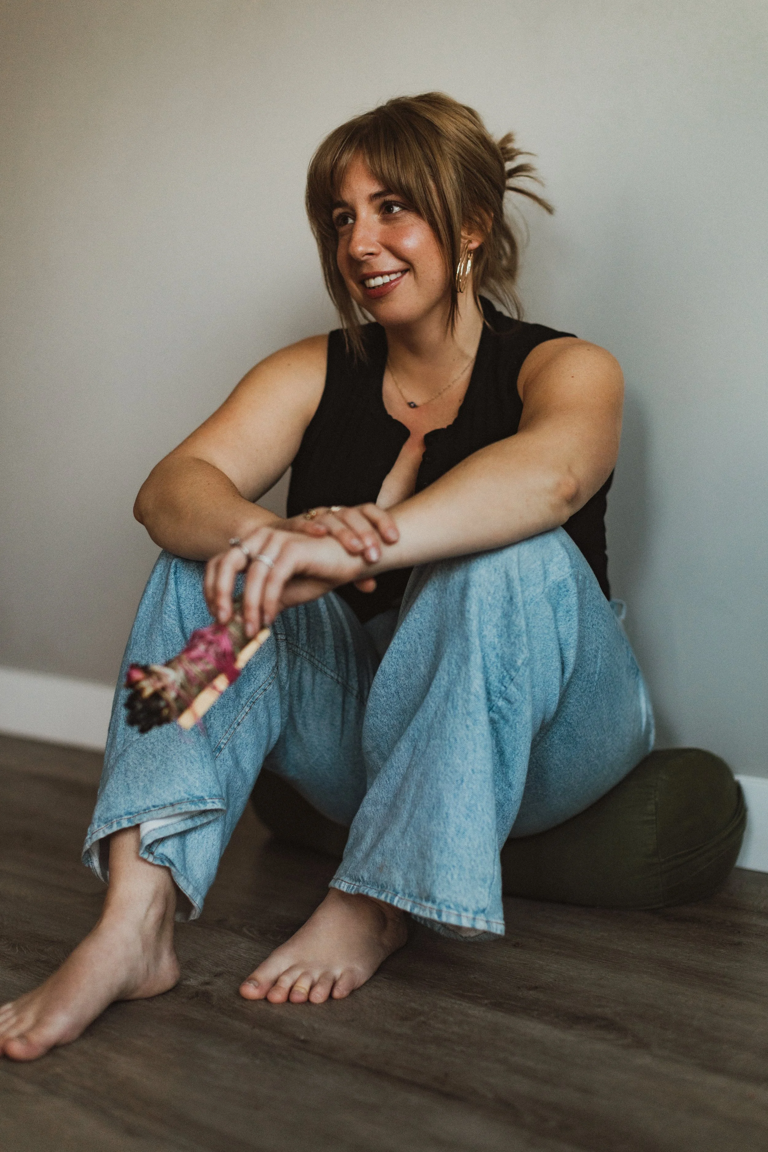 A woman with light brown hair sitting on the floor against a wall, smiling, holding a handmade object, wearing a black sleeveless top and loose blue jeans.