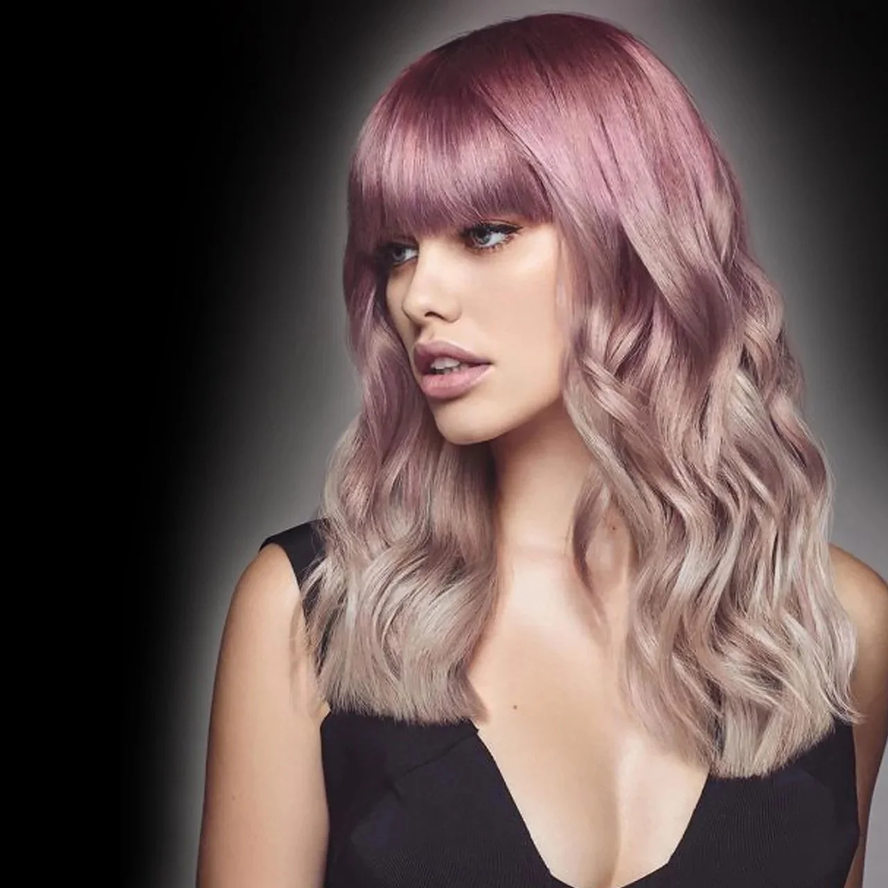 A woman with long wavy dyed pink hair and bangs, wearing a black sleeveless top, looking to the side against a dark background.