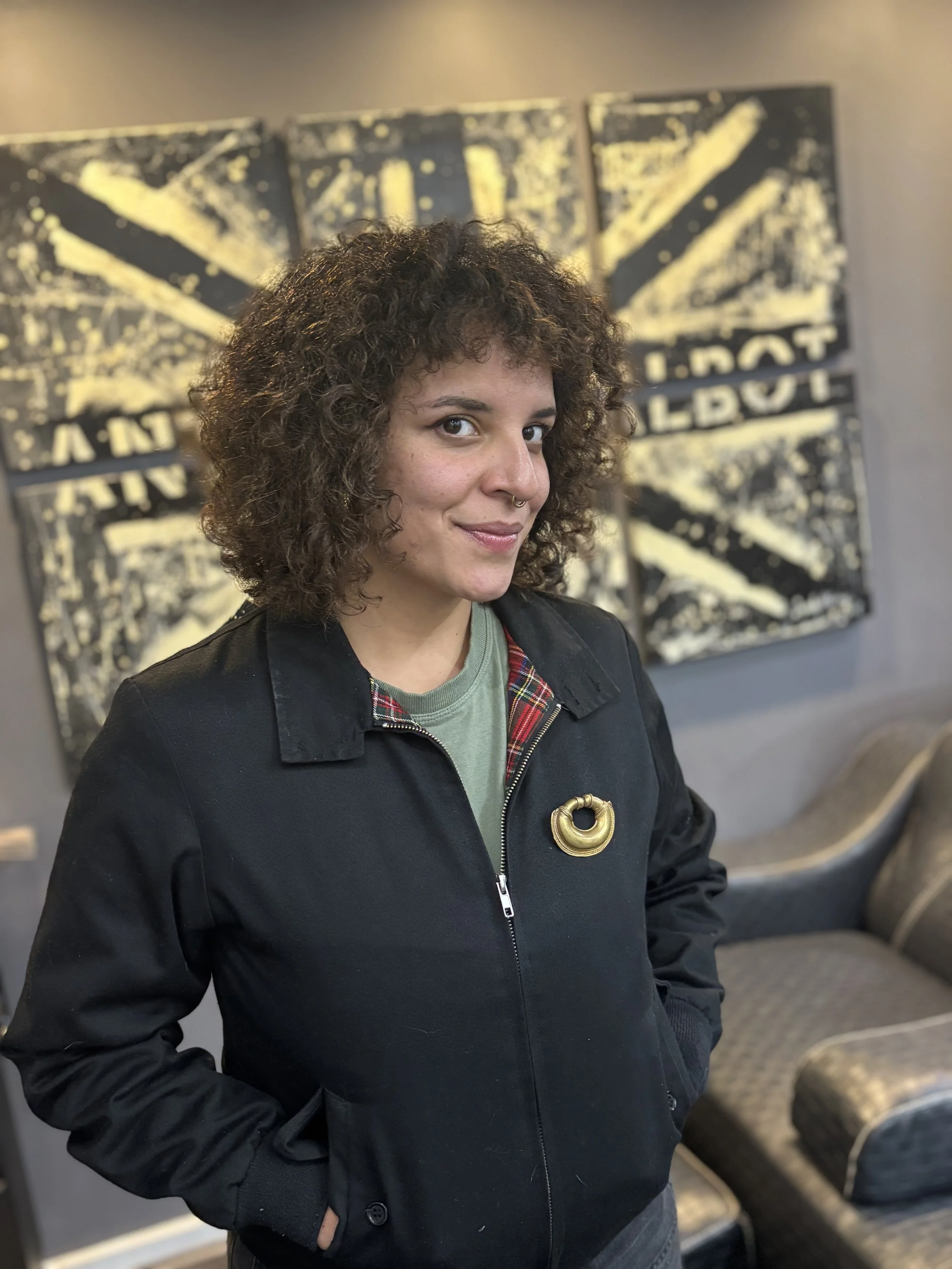 A woman with curly hair and a septum piercing standing indoors in front of an abstract black and yellow artwork. She is wearing a black jacket with a plaid lining and has her hands in her pockets.