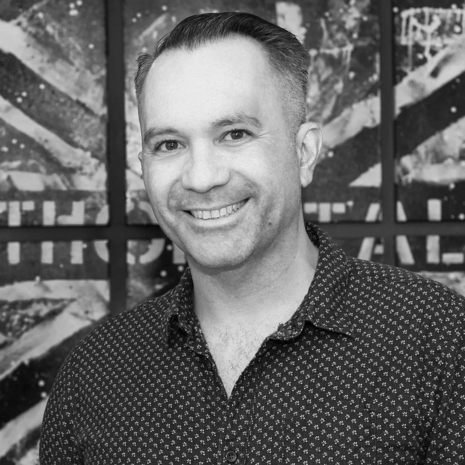 Black and white photograph of a smiling man with short, styled hair in front of a geometric abstract art background.