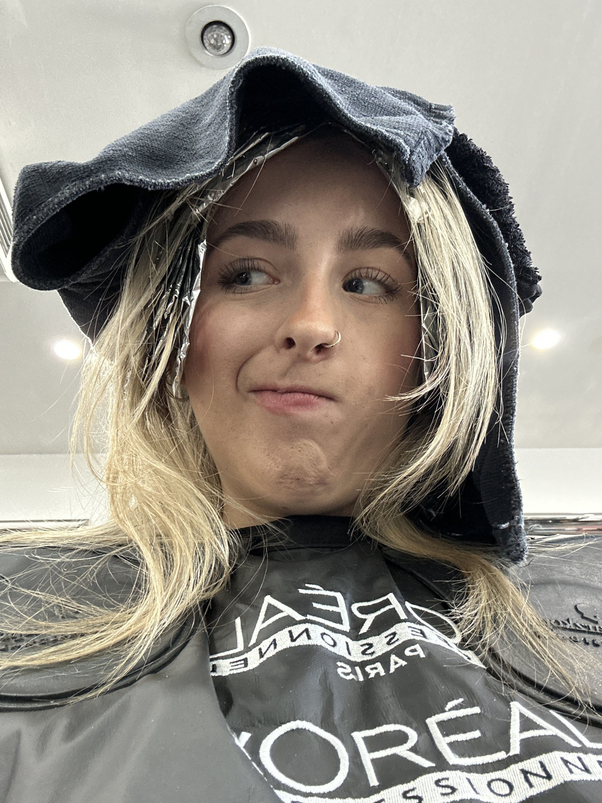 A young woman with blonde hair and a nose ring is in a salon or barbershop, wearing a L'Oréal Professionnel apron and a hoodie placed on her head while getting a haircut or styling.