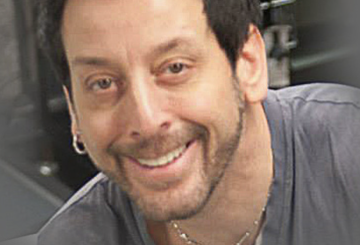 A smiling man with short dark hair, light skin, and a beard, wearing a gray shirt and a necklace, in an indoor environment.