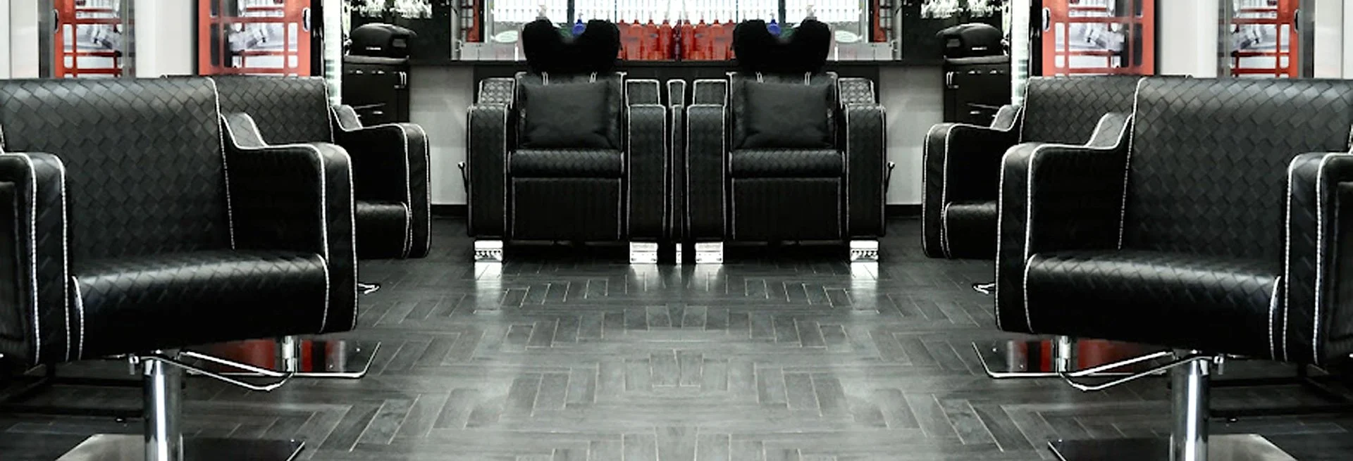 A symmetrical salon with black leather chairs and a dark wood floor. There are mirrors, styling tools, and red accents in the background.