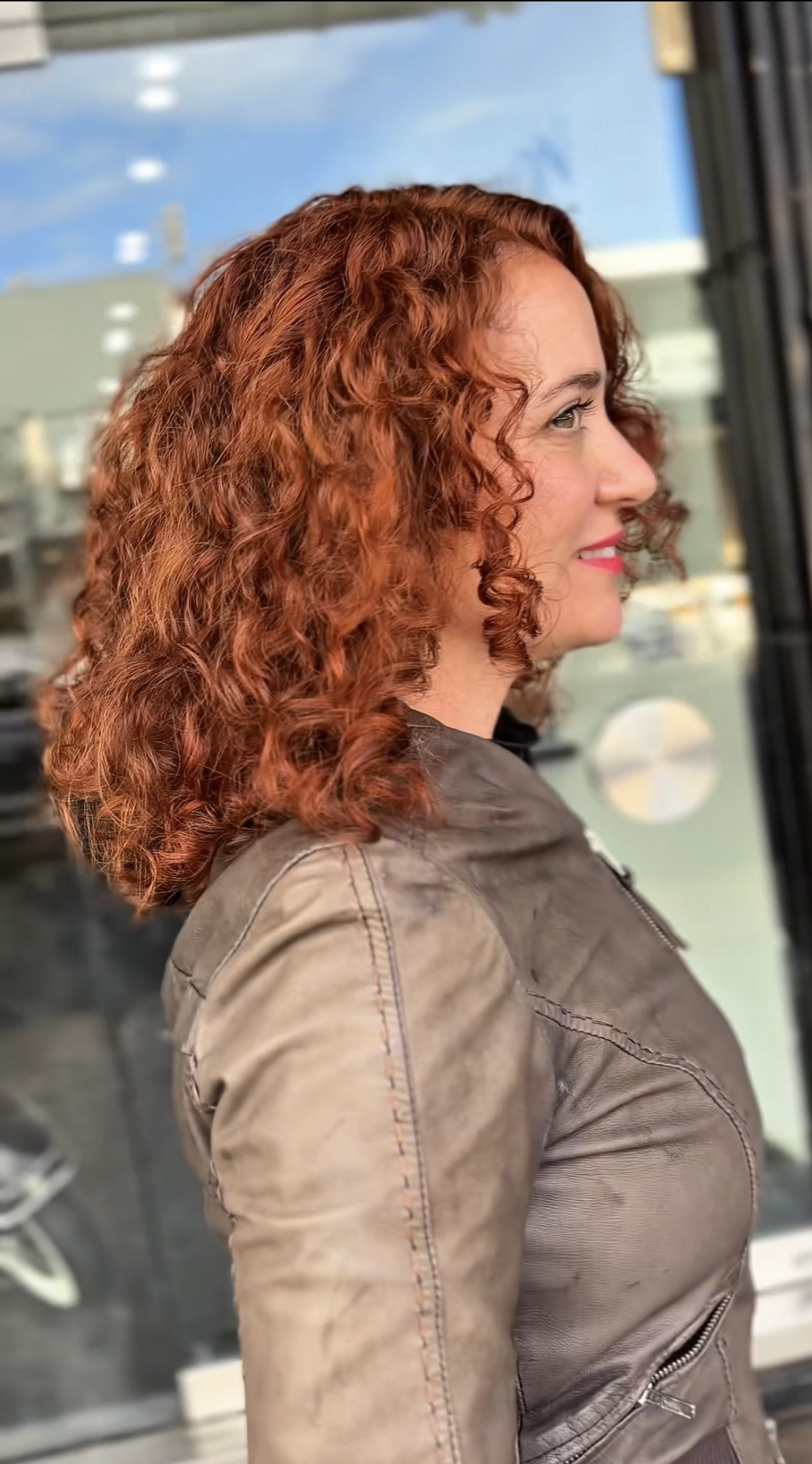 Side profile of a woman with curly red hair, smiling, wearing a brown leather jacket, standing outside near a glass window.