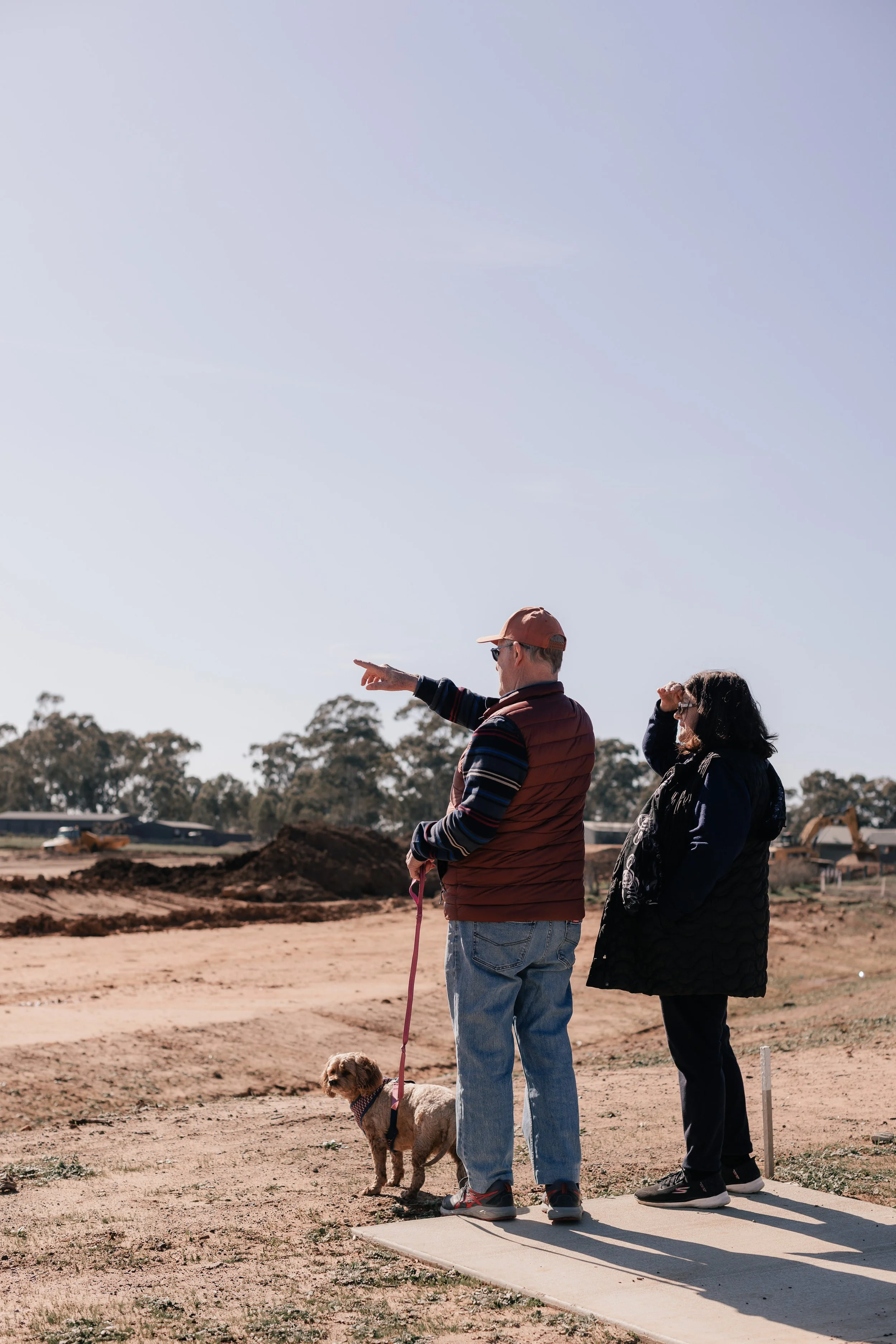 Two people and a dog standing on a construction site, with one person pointing towards the distance, and the other holding the dog on a leash.