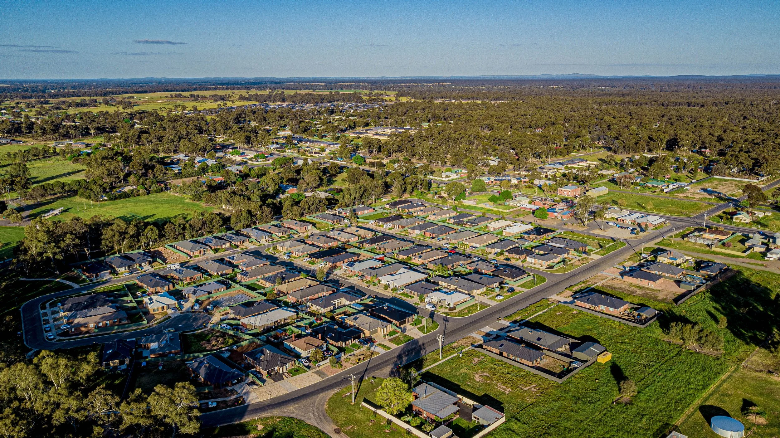 Aerial view of a suburban residential neighborhood with houses, streets, and green areas, surrounded by trees and open land.