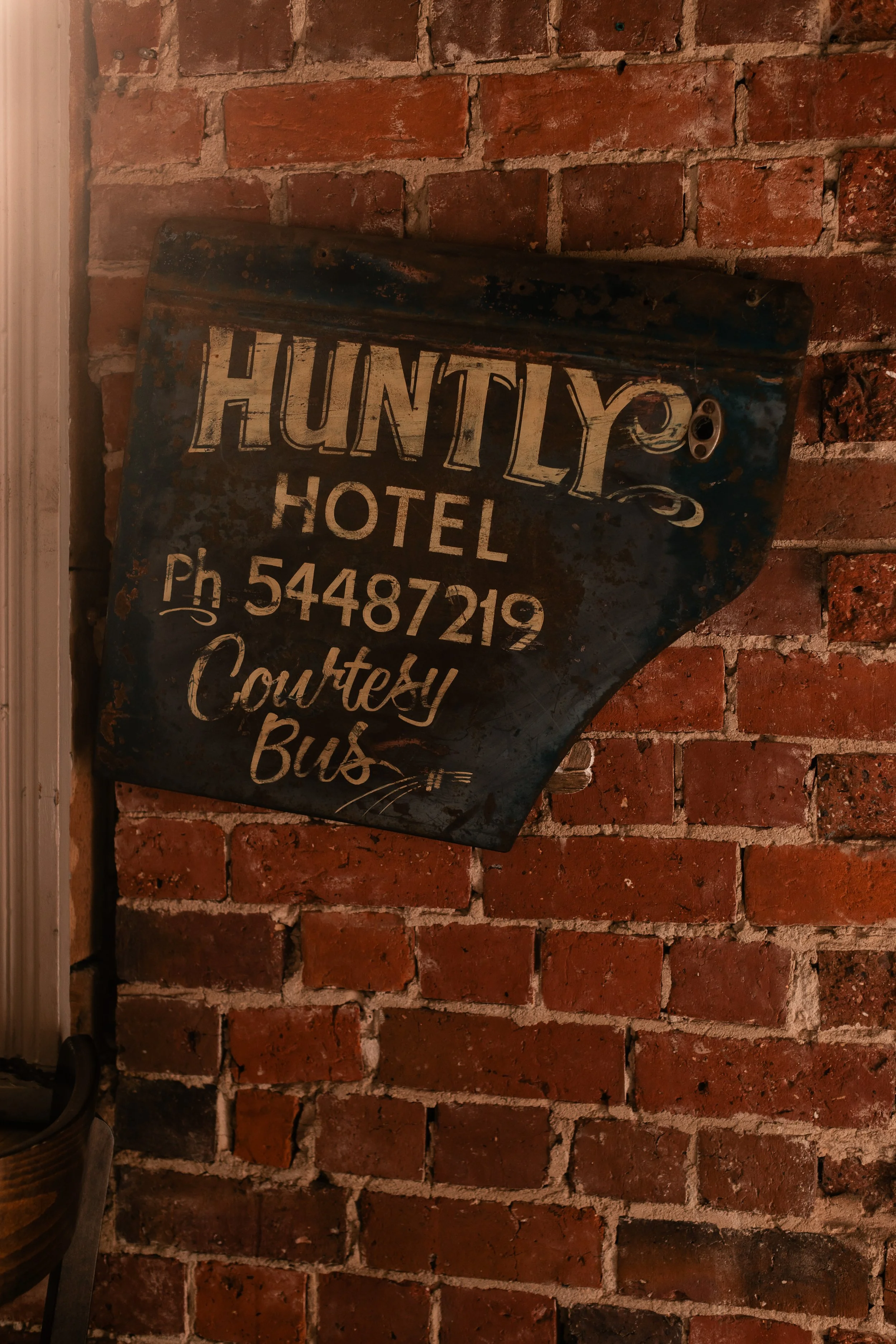 Vintage metal sign for Huntly Hotel mounted on exposed brick wall, includes phone number and indicates 'courtesy bus' service.