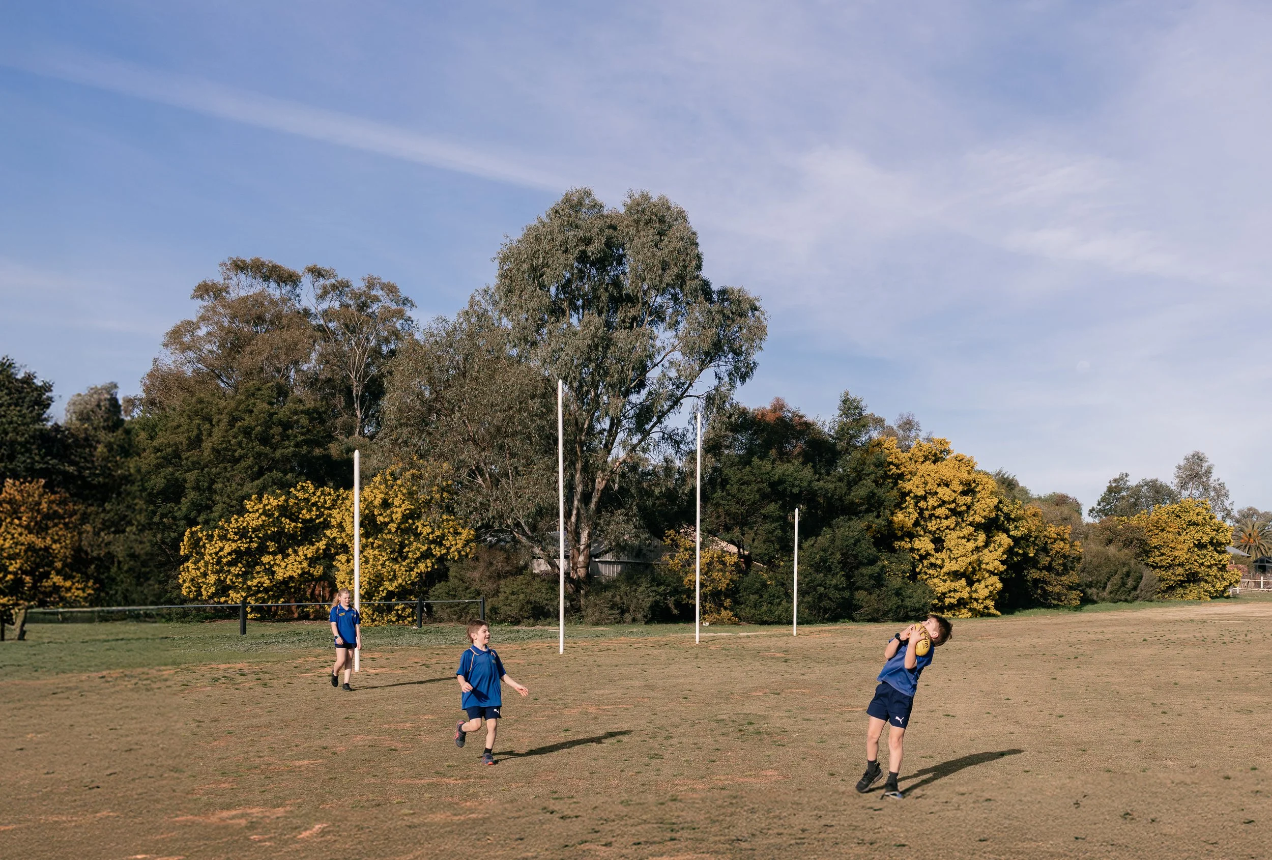 Three children in blue sports uniforms playing rugby on a dry field with goal posts, trees, and a blue sky in the background.