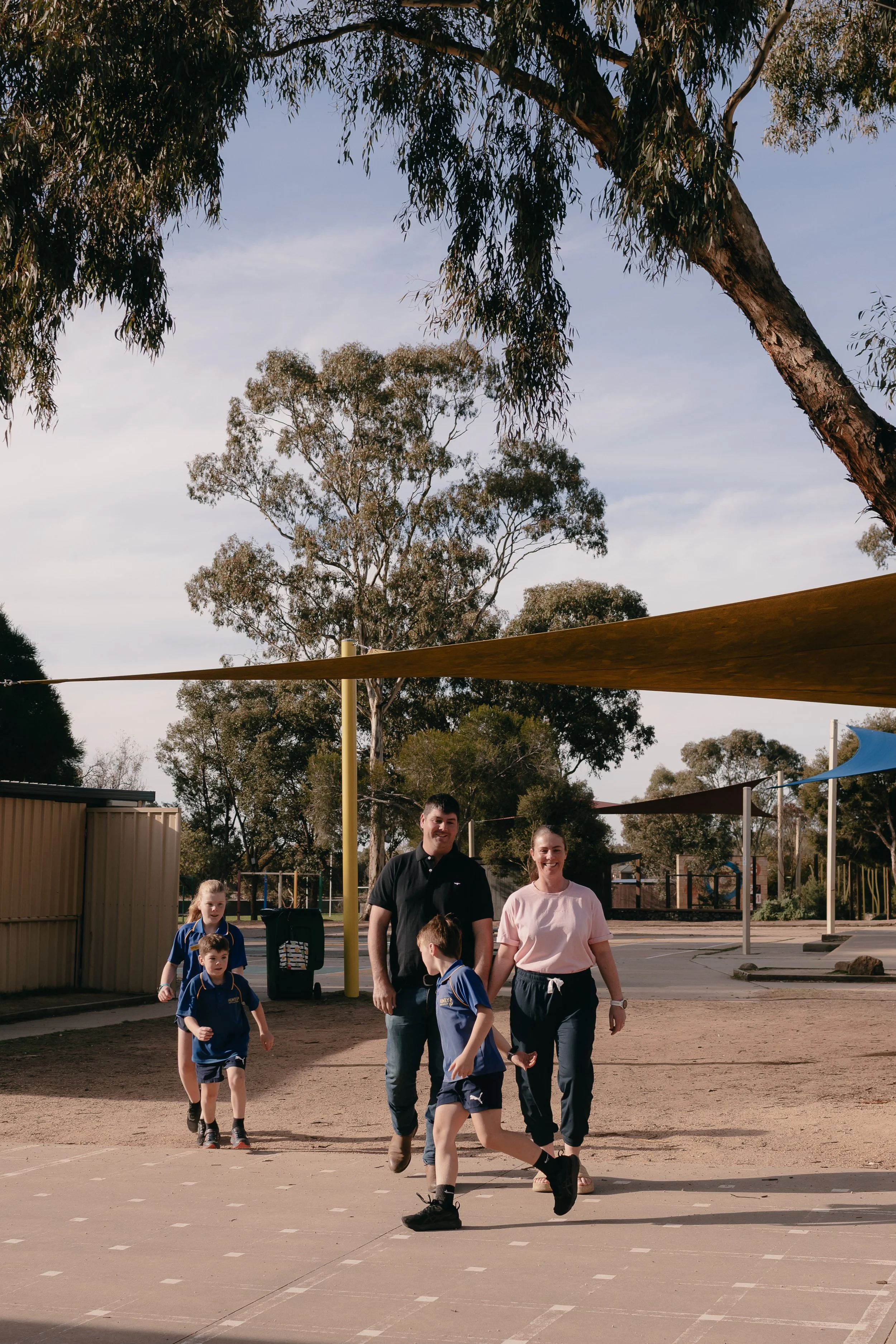 A group of children and adults walking in an outdoor playground area under shade sails, with trees and playground equipment in the background.
