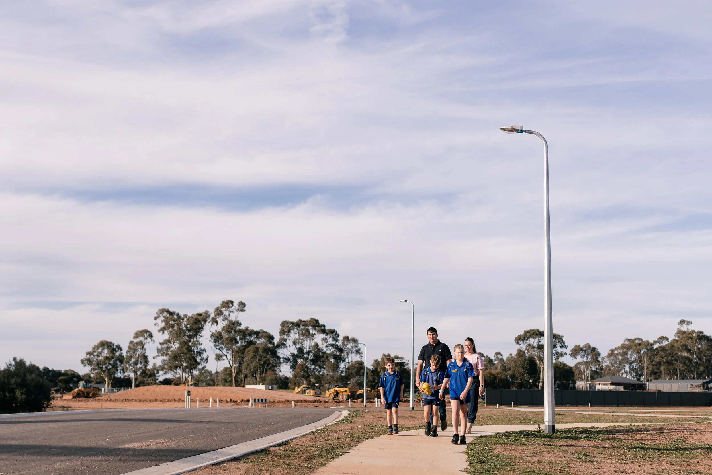 A group of children and an adult walking along a sidewalk next to a road, with tall streetlights and trees in the background under a partly cloudy sky.