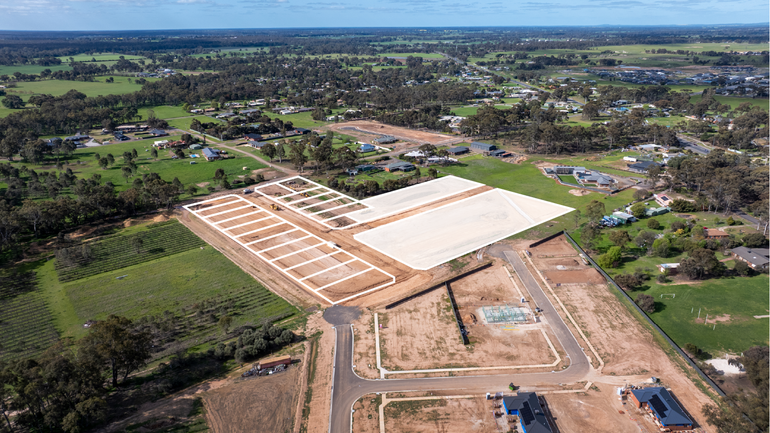 Aerial view of a construction site in a suburban area with multiple foundation slabs and cleared land, surrounded by green fields, trees, and residential houses.