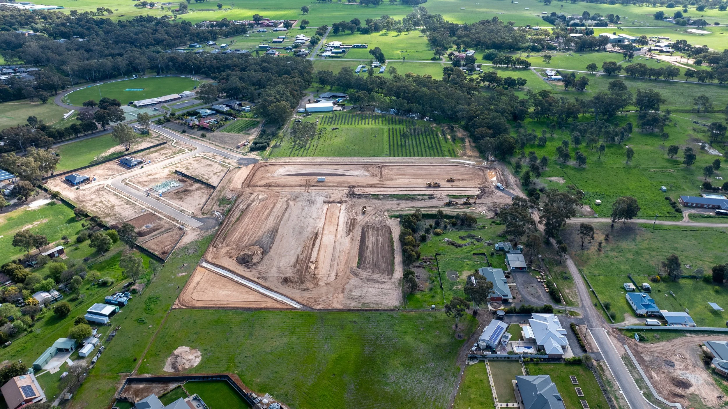 Aerial view of a construction site surrounded by residential houses, green fields, and trees. The site has cleared land, construction equipment, and dirt paths.