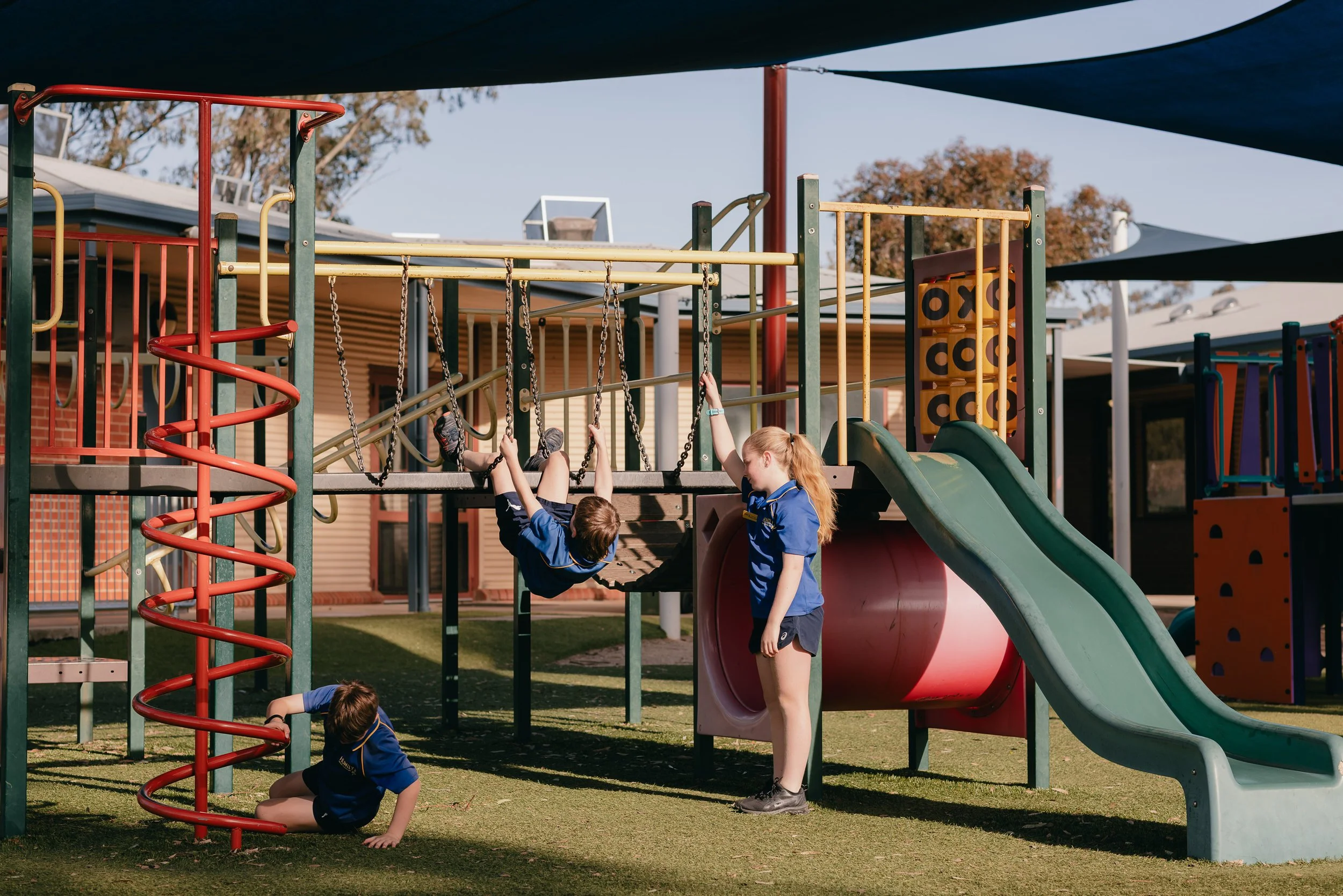 Children playing on a playground with slides, climbing structures, and swings, supervised by a girl in a blue shirt.