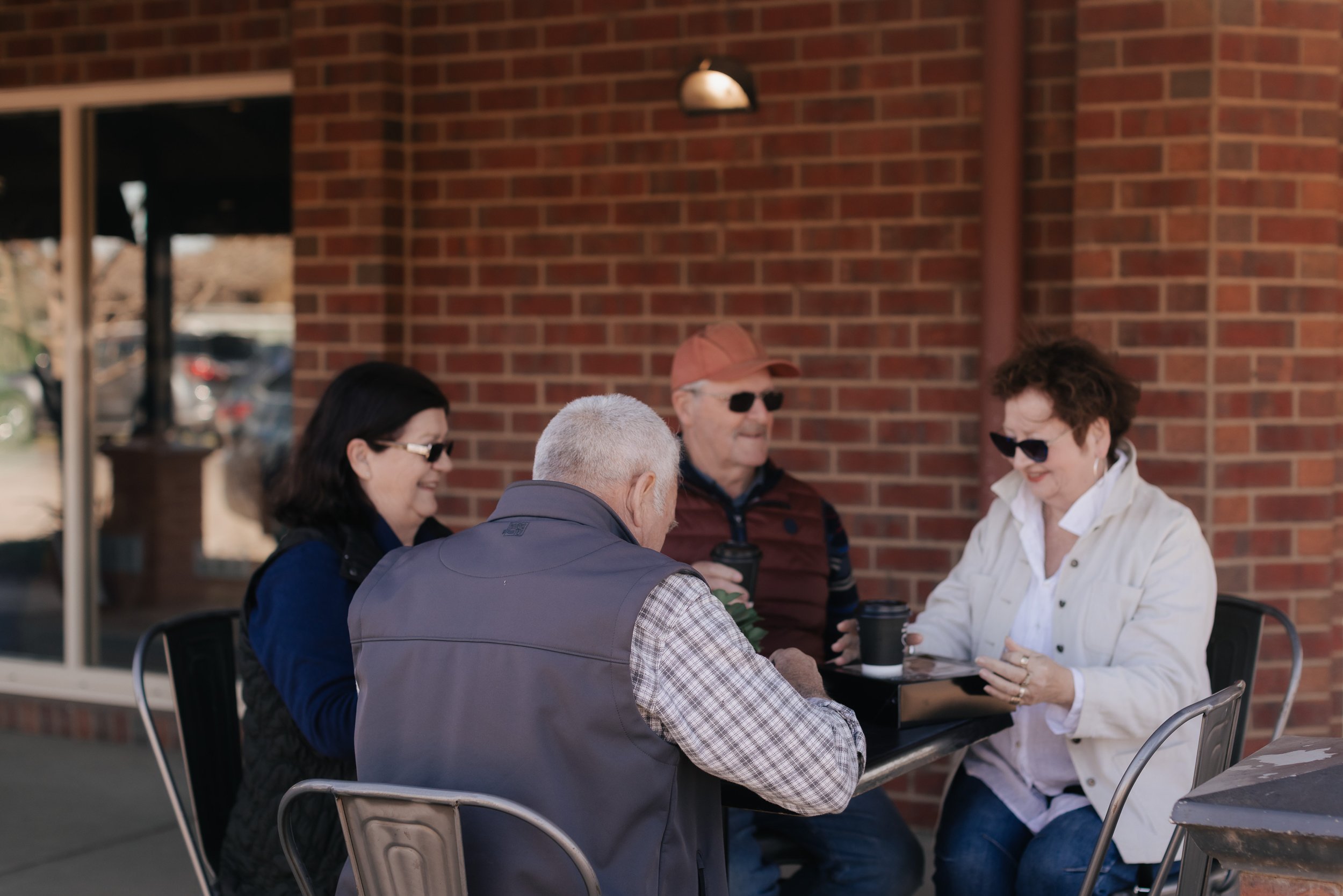 Four older adults sit at a table outside, enjoying coffee and conversation, with a brick building in the background.