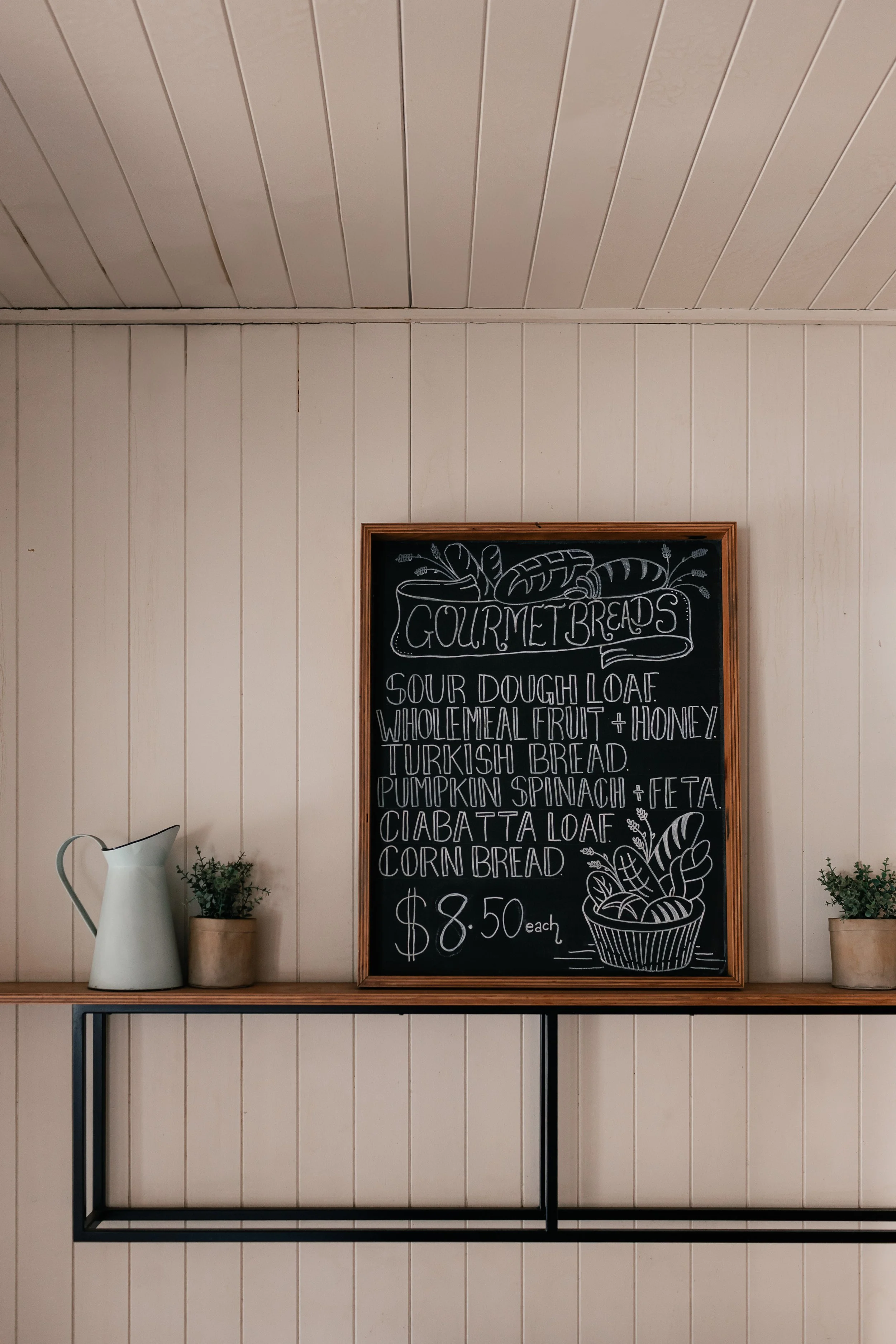 A chalkboard sign displaying a menu of gourmet breads, including sourdough loaf, wholemeal fruit and honey, Turkish bread, pumpkin spinach and feta, ciabatta loaf, and corn bread, priced at $8.50 each, situated on a wooden shelf with a watering can a