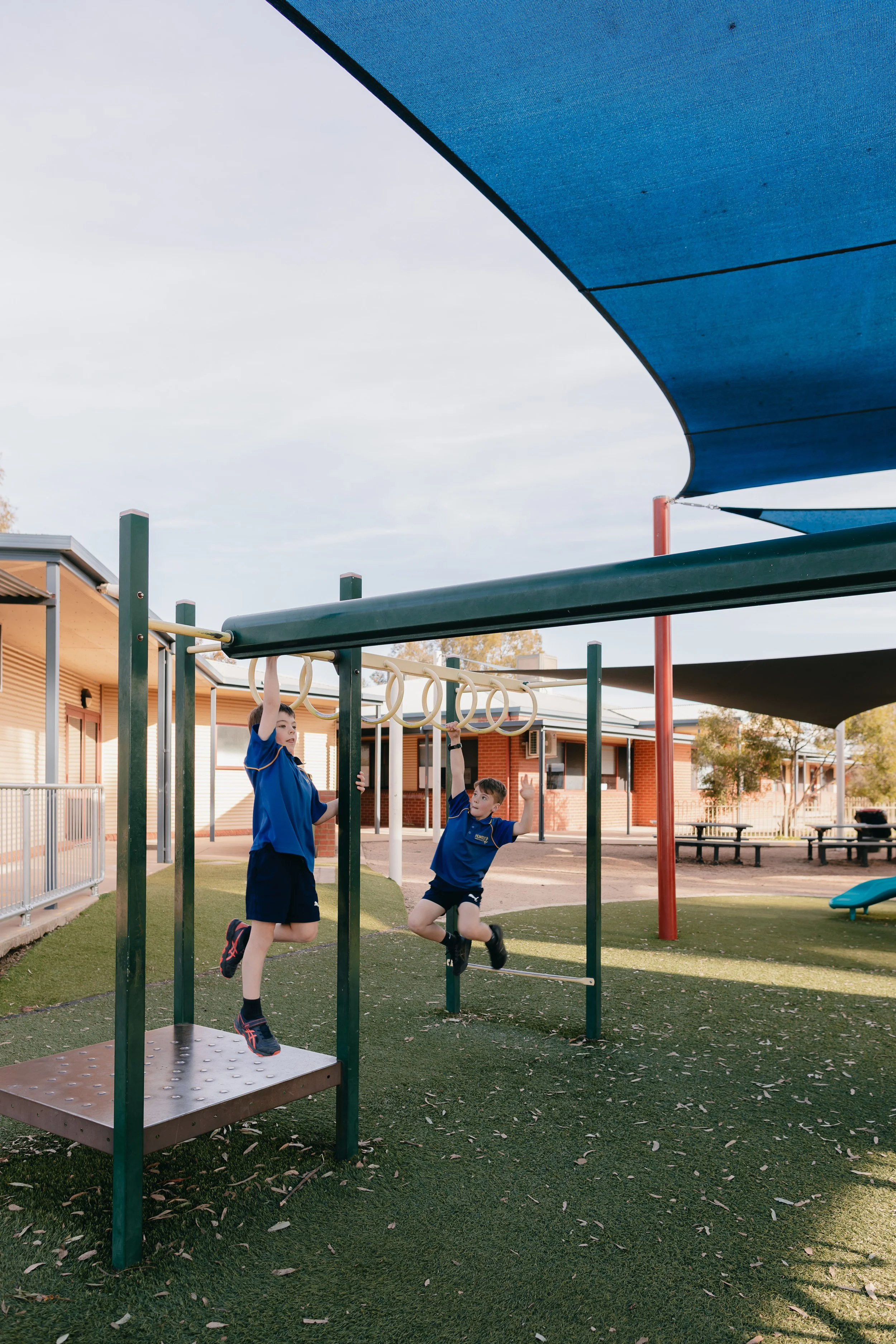 Two children playing on gymnastic rings at a school playground with shade sails overhead.