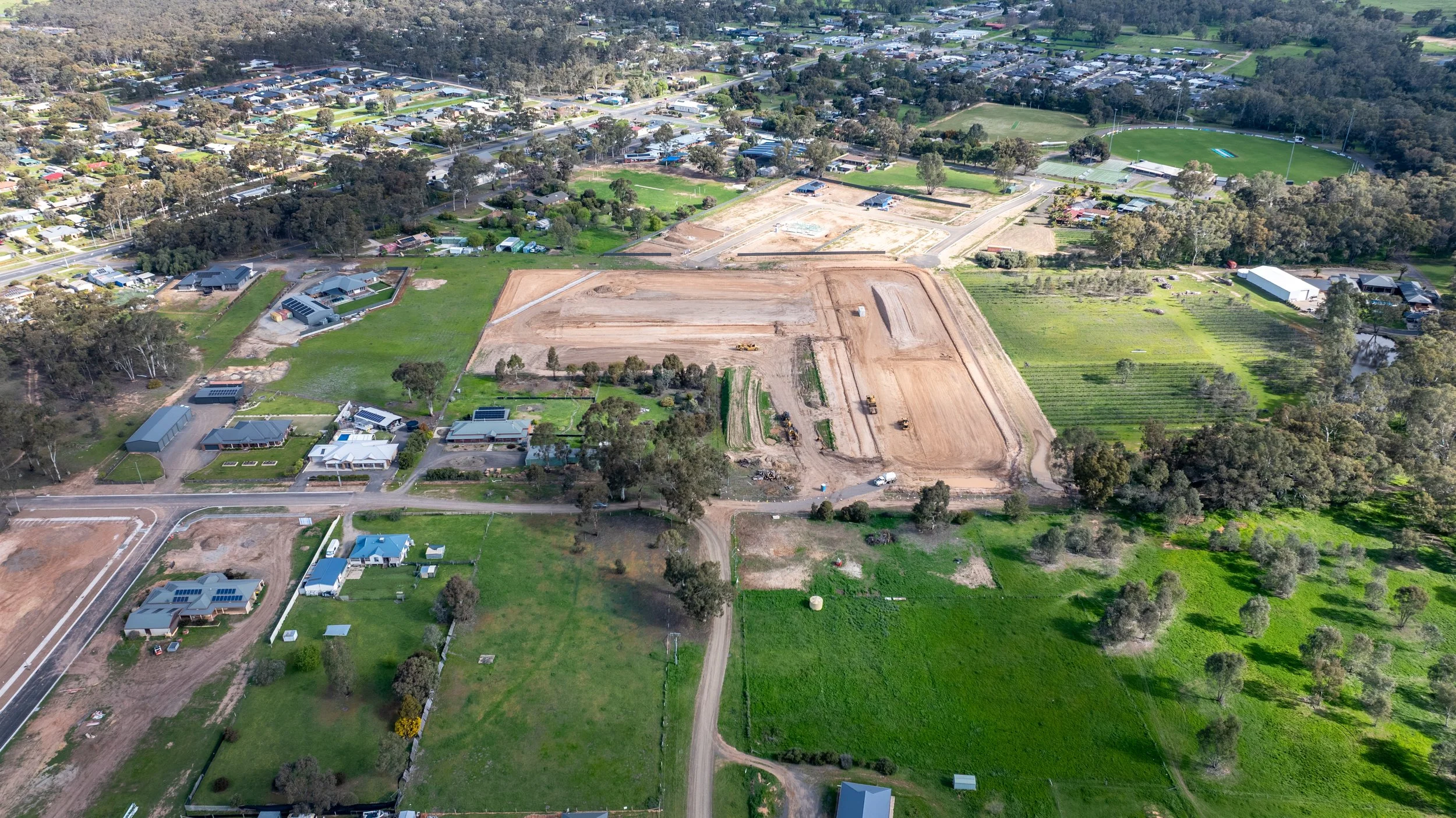 Aerial view of a residential area with houses, roads, and a large construction site with earth-moving equipment amidst greenery.