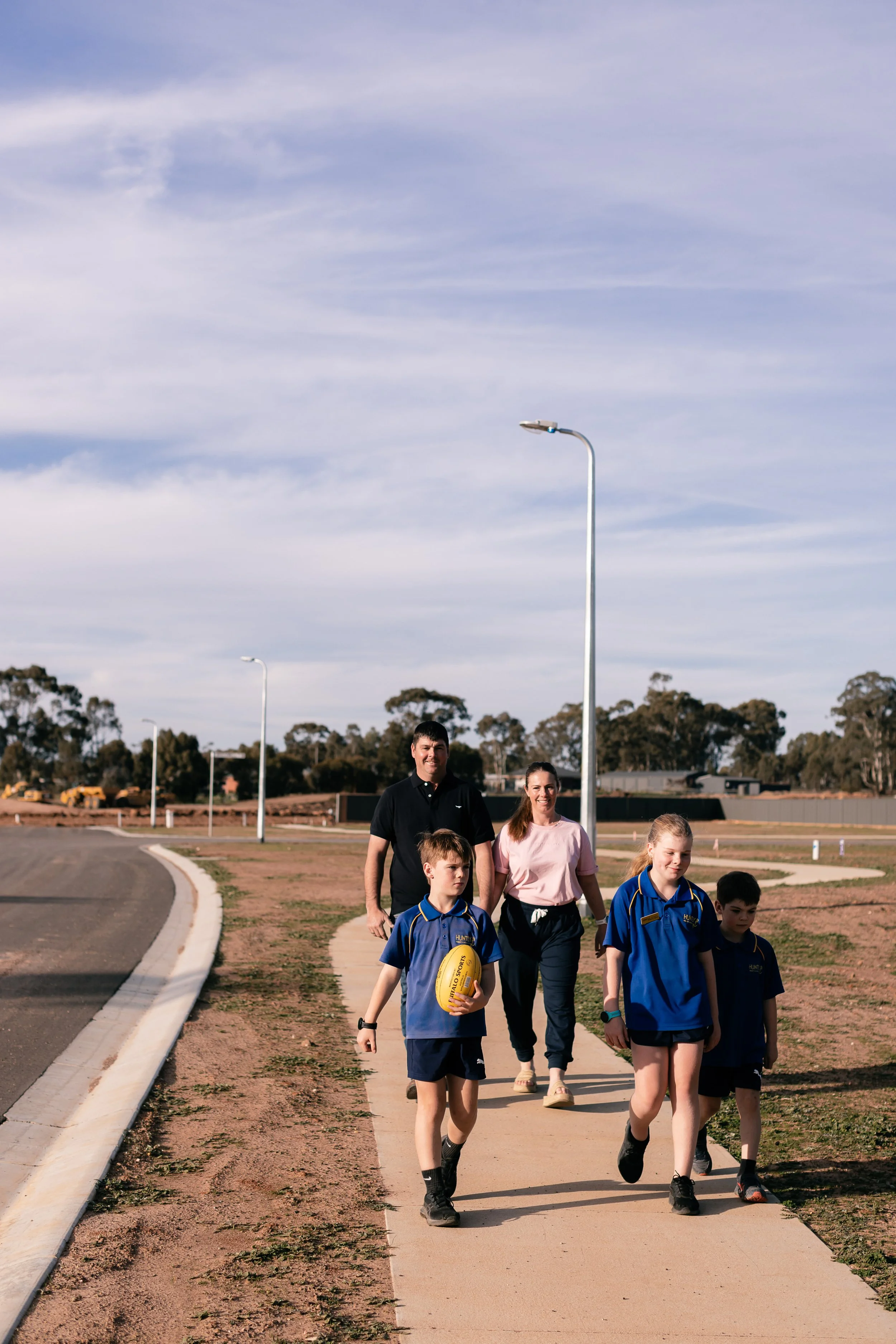 A group of children and two adults walking on a sidewalk in an open outdoor area on a cloudy day. The children are wearing school uniforms, and one boy is holding a yellow football.