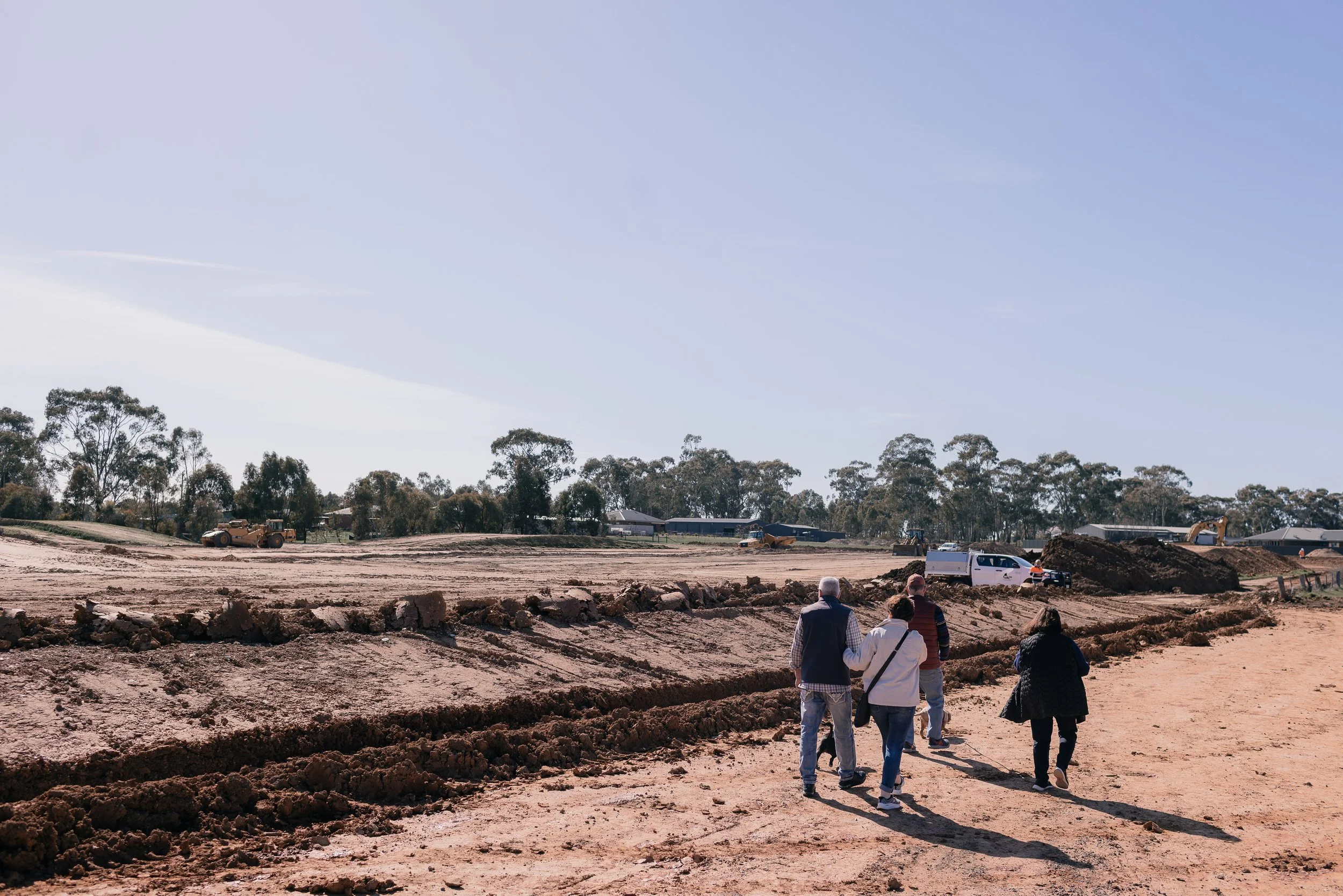 Four people walking on a dirt path at a construction site with heavy machinery and excavated earth, and trees in the background.